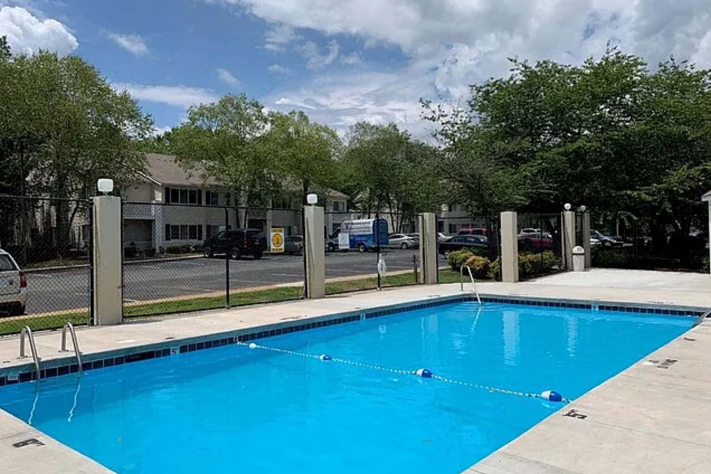 A bright blue swimming pool surrounded by a concrete patio, with safety lanes marked by floating buoys. Lush green trees and residential buildings are visible in the background, under a partly cloudy sky. Fences and light poles line the area, creating a relaxing atmosphere.
