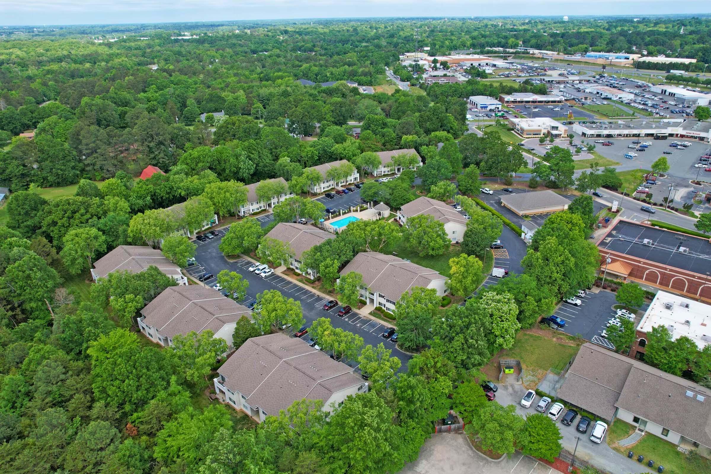 Aerial view of a residential complex surrounded by lush greenery, featuring several buildings arranged in a circular layout with a central swimming pool. Nearby, a commercial area with various stores can be seen in the background, highlighting a mix of nature and urban development.