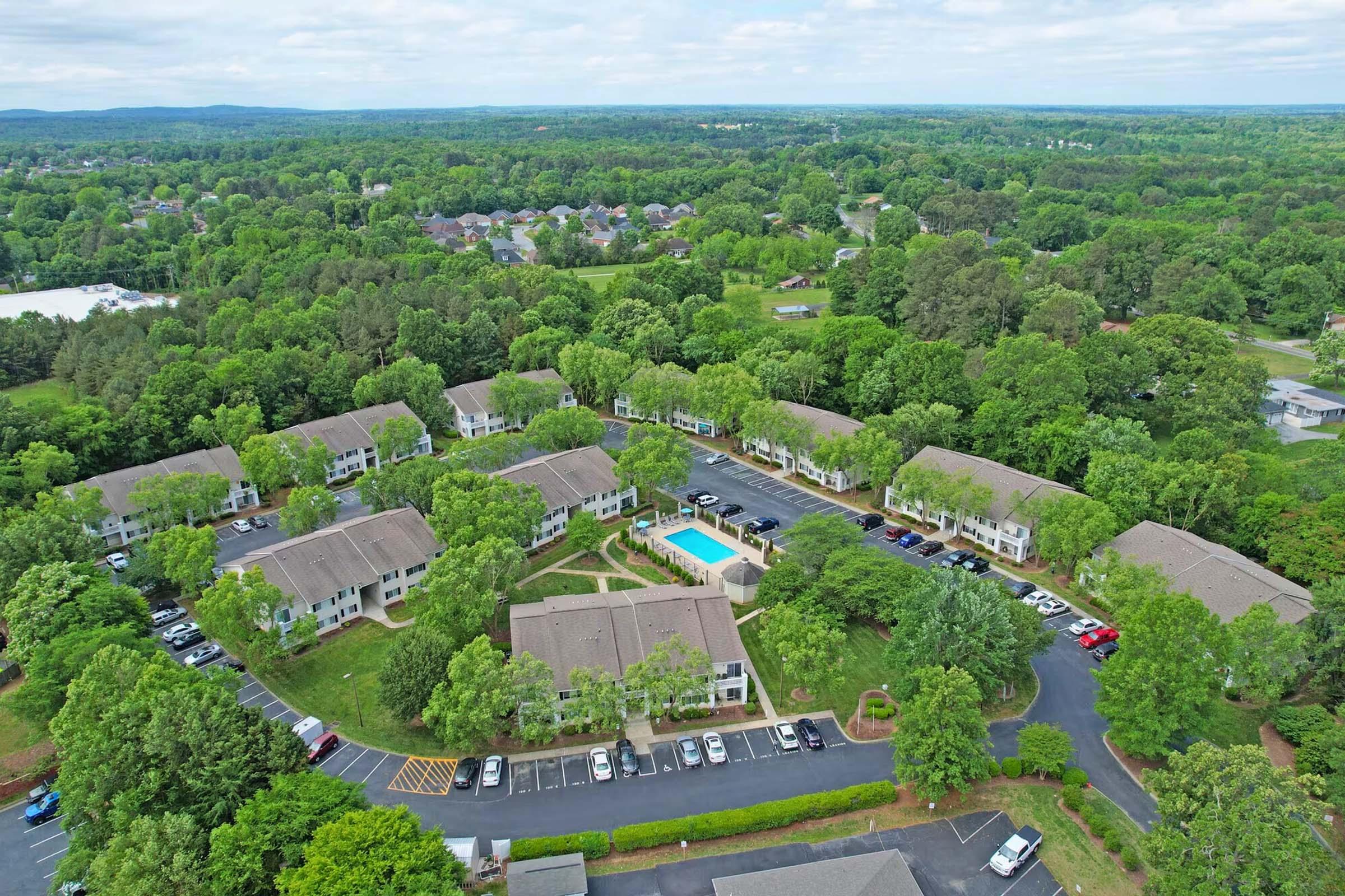 Aerial view of a residential community surrounded by greenery. The layout includes multiple buildings arranged in a circular pattern, with a central swimming pool and ample parking space. The landscape features trees and lush fields, creating a serene environment. Cloudy sky above.