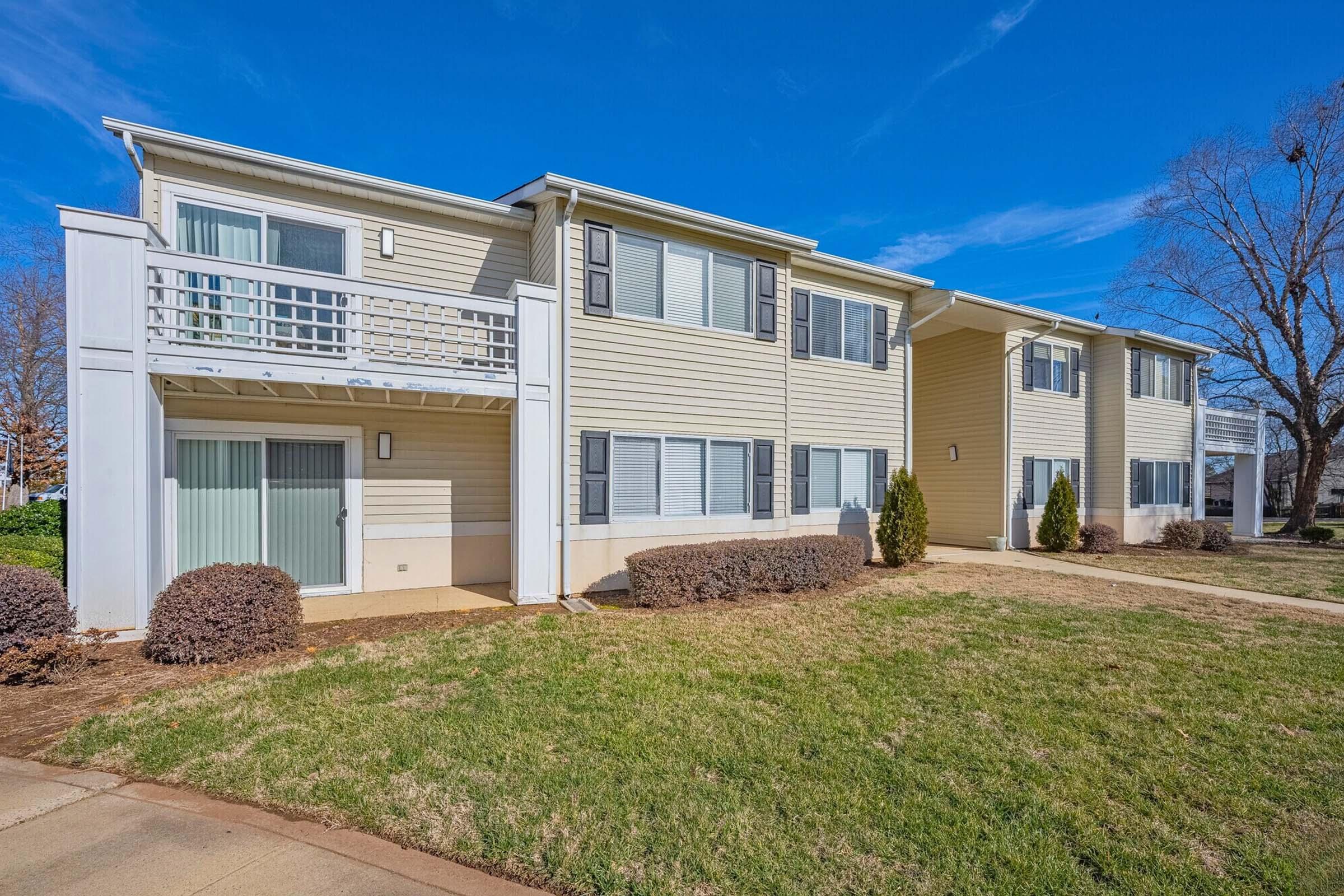 A two-story residential building with light yellow siding and white trim. The facade features multiple windows and balcony spaces. The surrounding area includes neatly trimmed grass and small shrubs, with a clear blue sky in the background.
