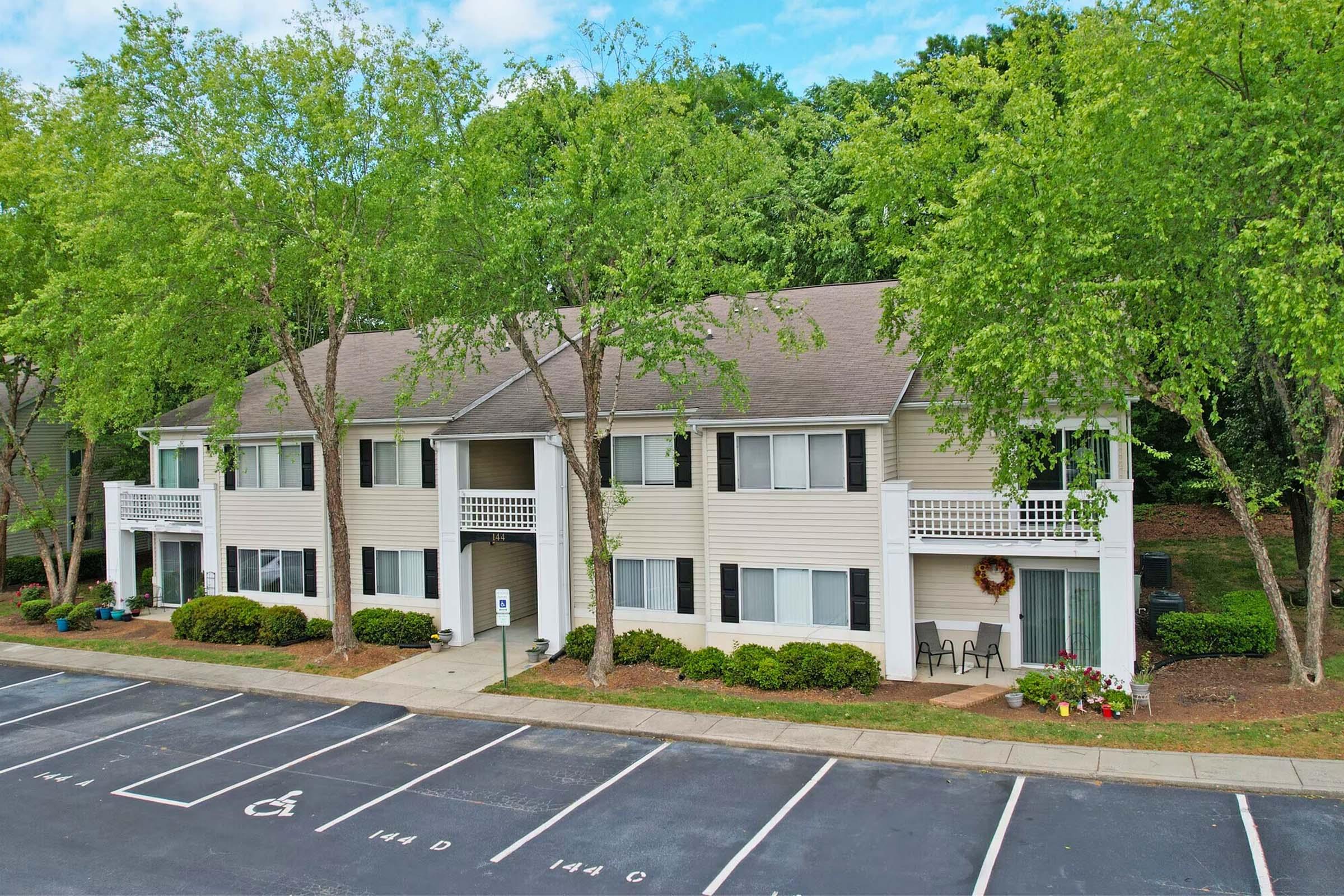 A two-story apartment building surrounded by greenery, featuring balconies with chairs. The building has a light-colored exterior and a welcoming entrance. Parking spaces are visible in the foreground, labeled with numbers. The scene is bright and inviting, showcasing a peaceful residential area.