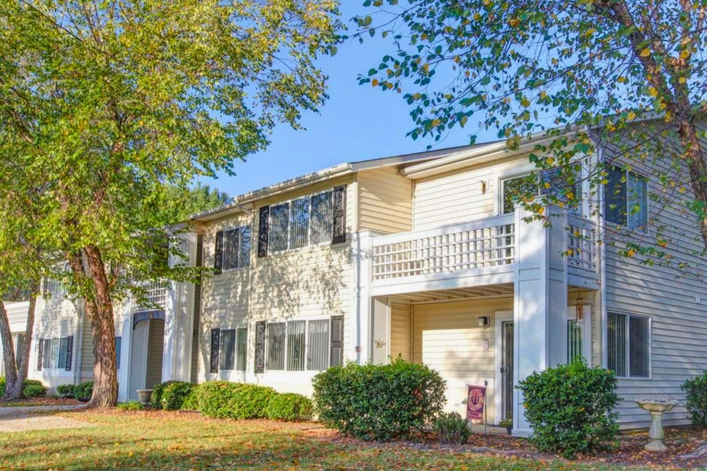 A two-story beige apartment building with multiple windows and balconies, surrounded by green shrubs and trees. The scene features a clear blue sky and autumn leaves on the ground, creating a bright and inviting atmosphere.