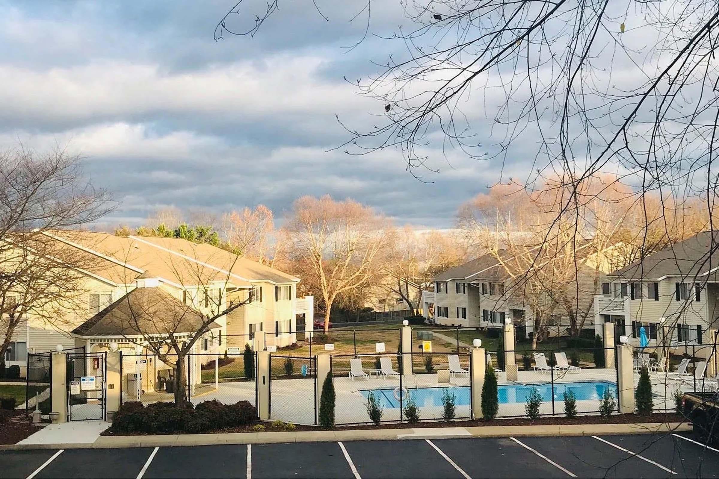 View of a residential apartment complex featuring a swimming pool surrounded by lounge chairs. Trees with sparse foliage are visible in the foreground, and a cloudy sky looms above. The scene is tranquil and inviting, suggesting a relaxed outdoor atmosphere.