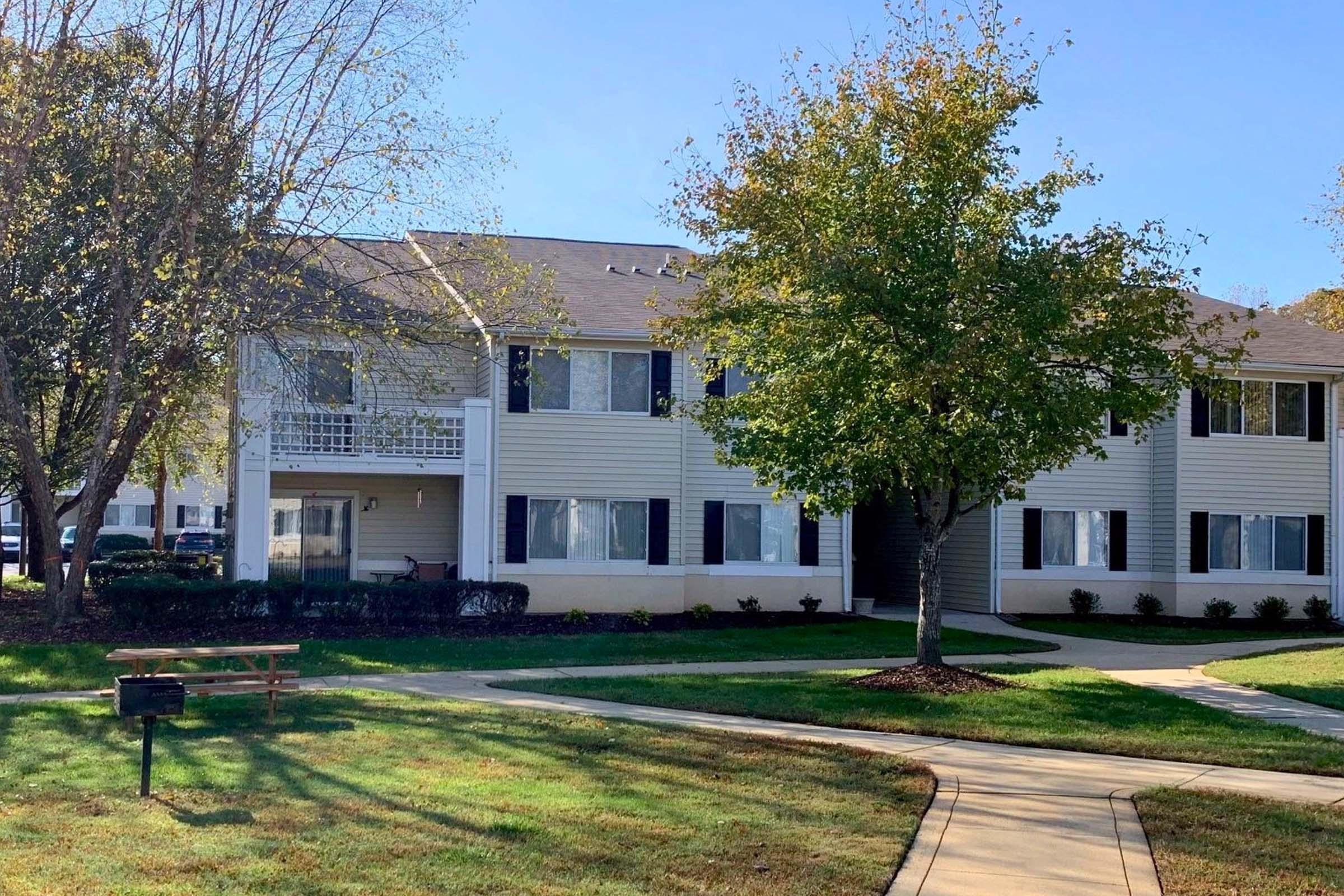 A two-story residential building with light-colored siding, featuring multiple windows and a balcony on the left side. In the foreground, there is a small picnic table and a tree, with well-maintained grass and landscaping surrounding the pathway leading to the entrance.