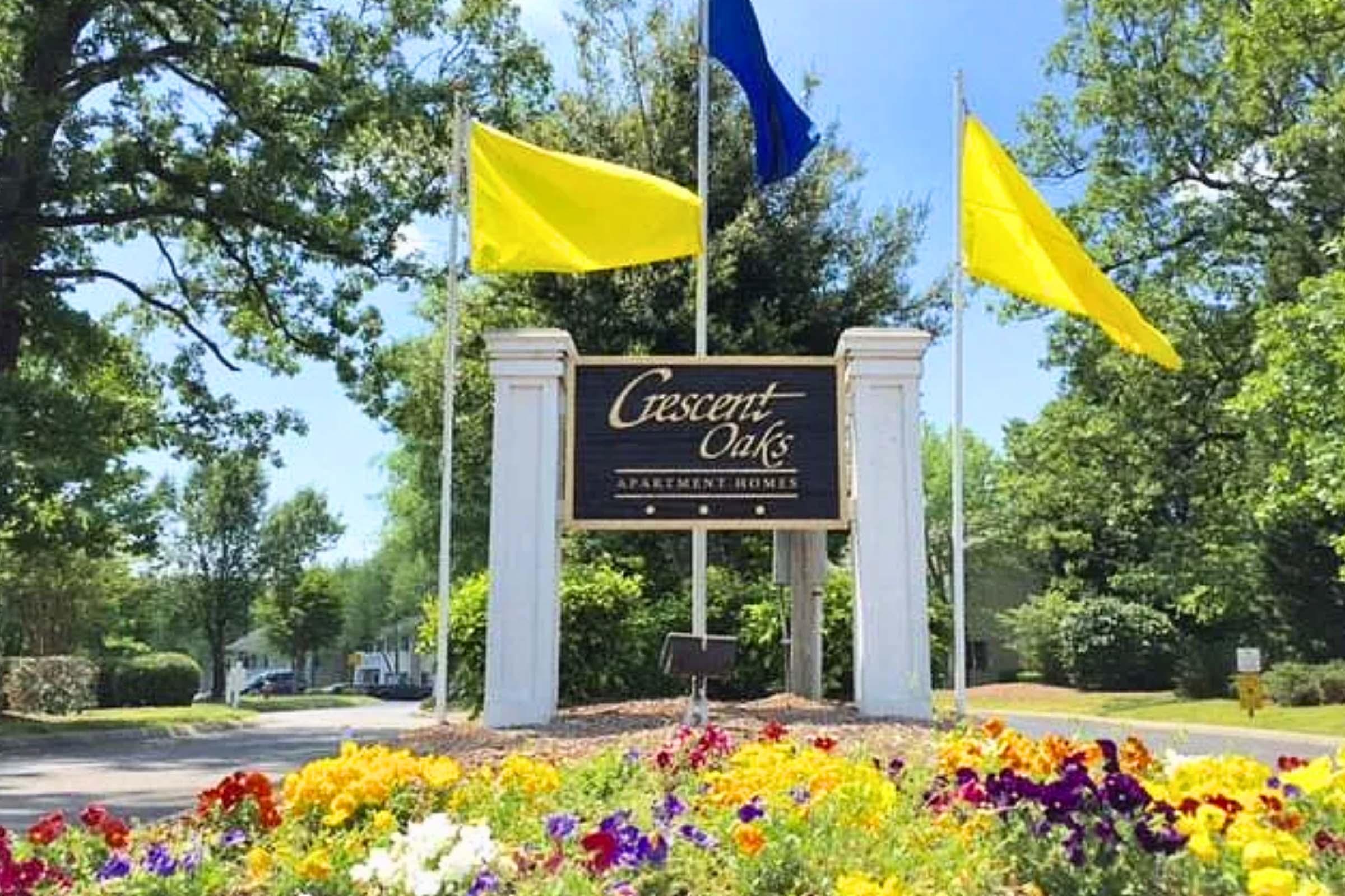 Sign for Crescent Oaks Apartment Homes, featuring yellow flags on either side. The entrance is surrounded by colorful flower beds, with trees in the background. The setting is bright and welcoming, conveying a pleasant atmosphere for residents and visitors.