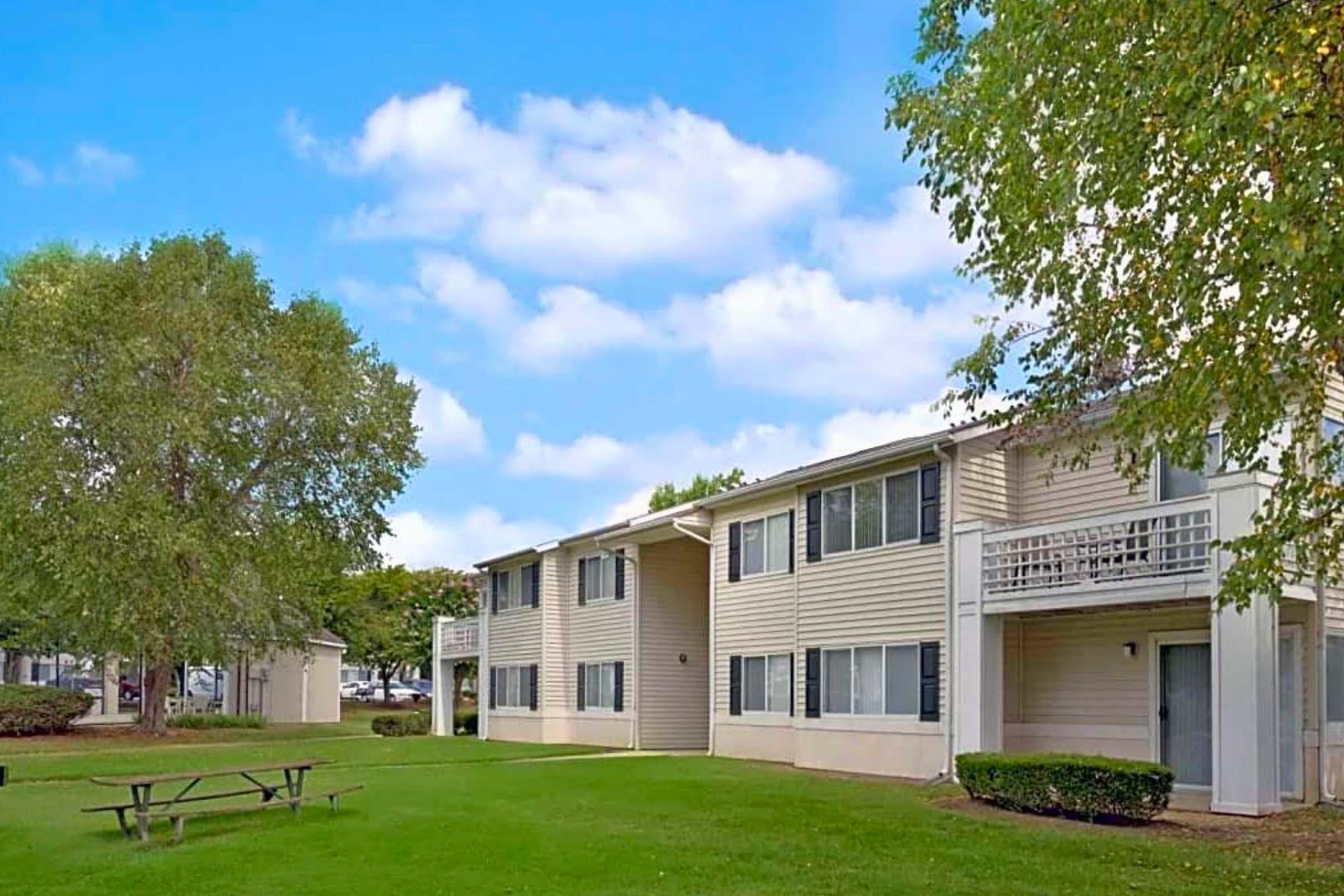 A two-story apartment building with light-colored siding, surrounded by a grassy area and trees. There is a picnic table in the foreground, and the sky is partly cloudy, creating a bright and inviting atmosphere.