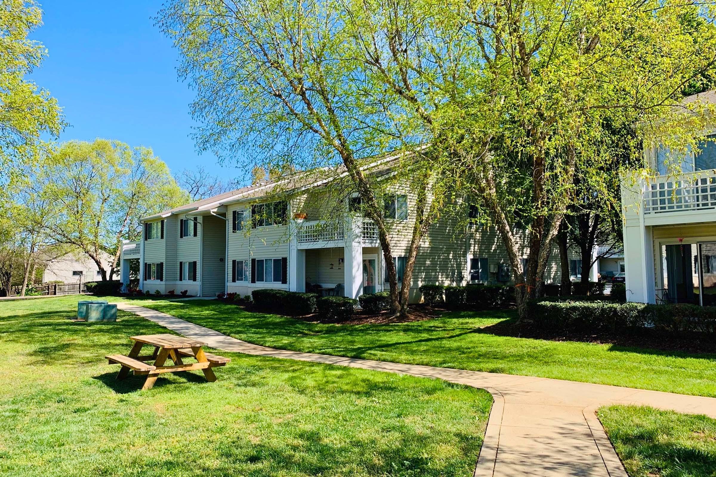 A sunny scene of a green lawn with a paved pathway leading to a two-story building surrounded by trees. A picnic table is placed on the grass in the foreground, adding to the inviting atmosphere of the outdoor space. Clear blue sky is visible above.