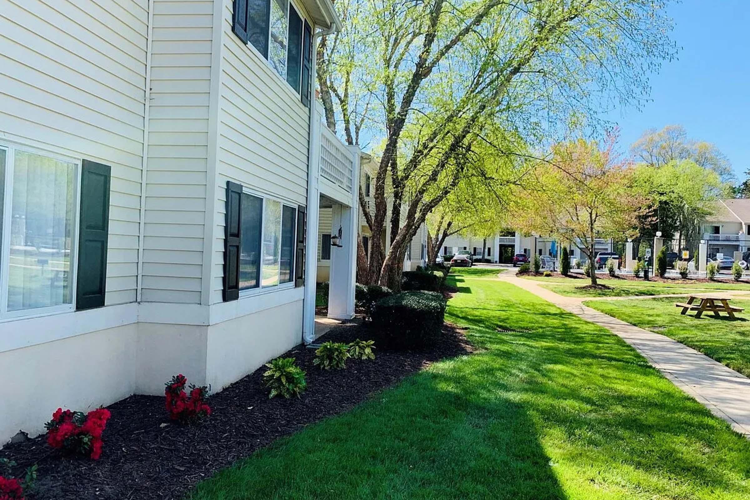 A well-maintained apartment complex surrounded by vibrant greenery, featuring a paved pathway, neatly trimmed bushes, and flowering plants. Sunlight filters through trees, indicating a pleasant and inviting outdoor environment. Bright blue skies add to the overall cheerful atmosphere.