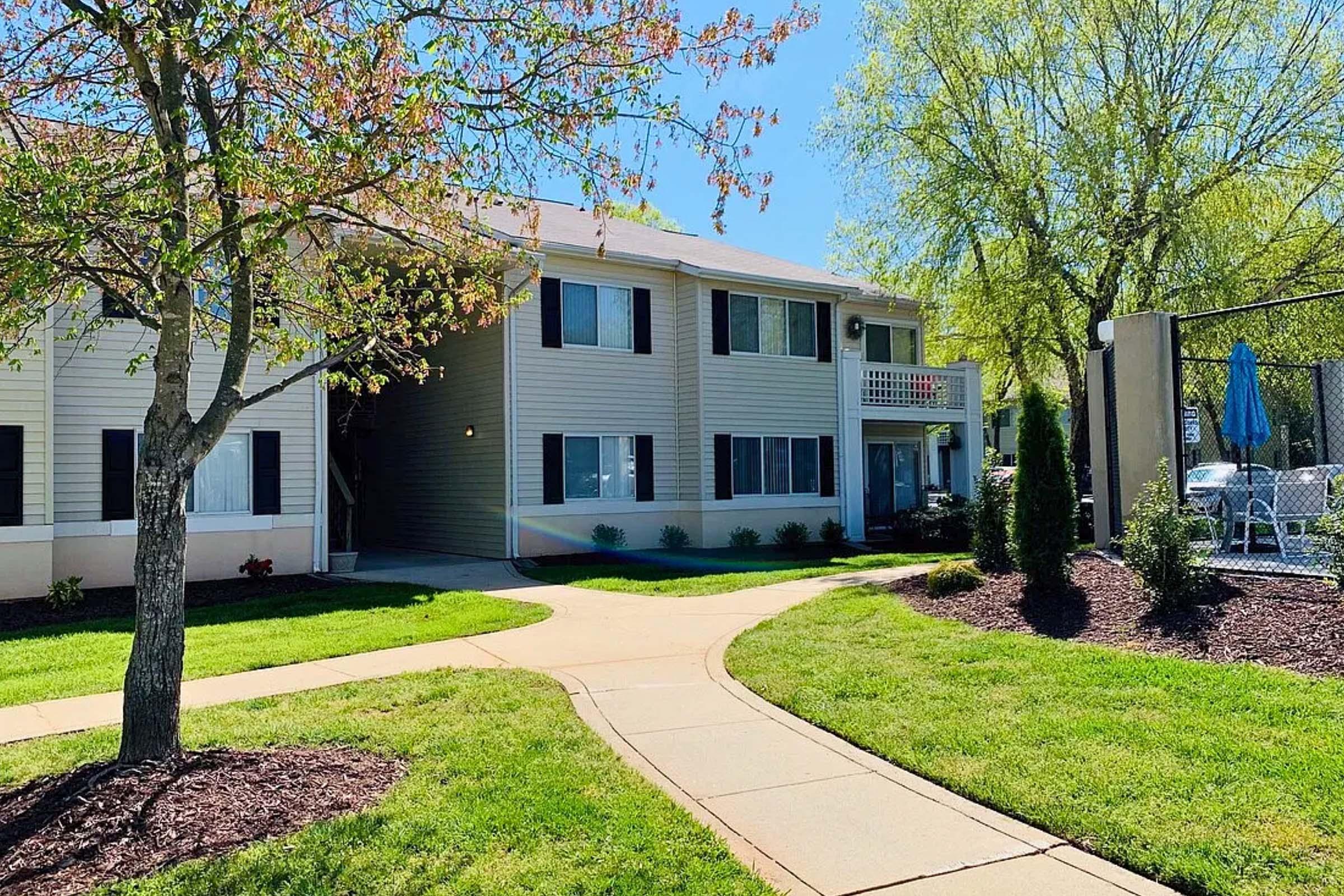 A bright and sunny view of a residential building with light-colored siding, surrounded by manicured lawns and blooming trees. A pathway winds around the building, and a fenced-in pool area is visible in the background, indicating a welcoming community atmosphere.