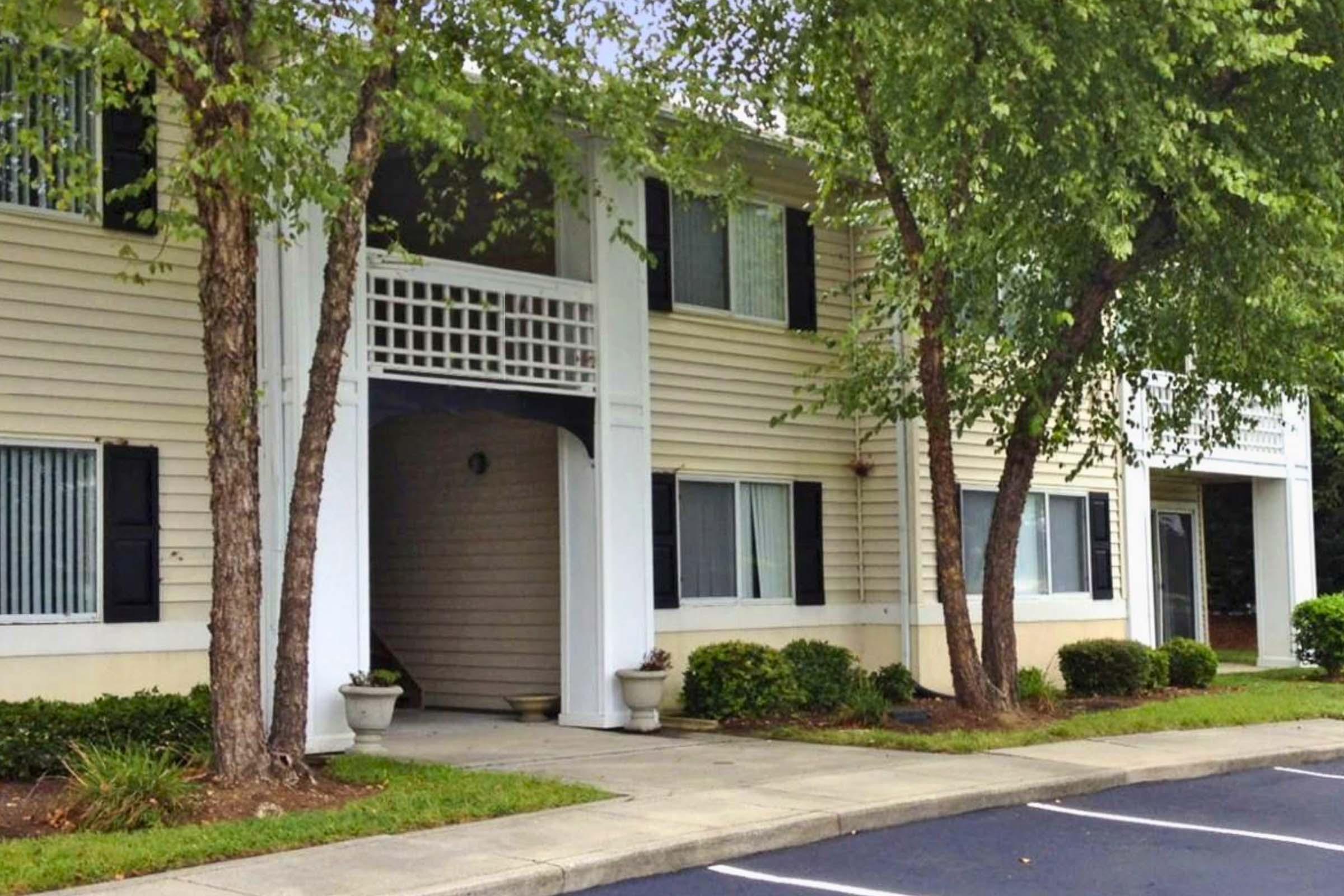 Exterior view of a residential building featuring a light-colored facade, symmetrical windows with black shutters, and a covered entrance. Lush greenery surrounds the entrance, and there is a parking lot in front with neatly trimmed shrubs along the walkway.