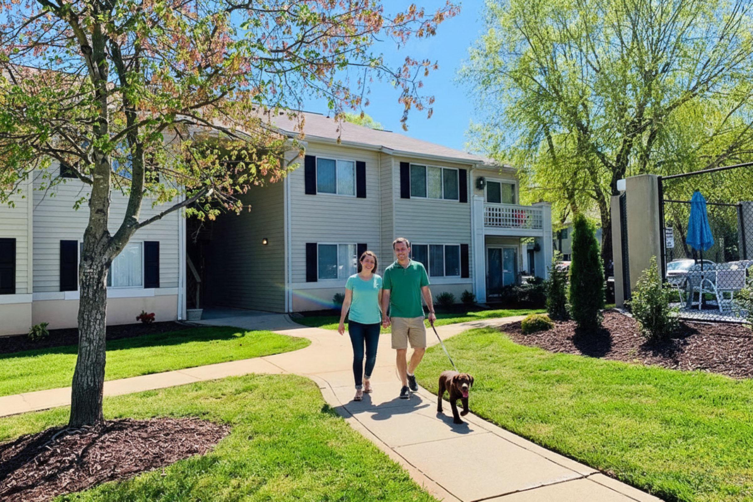 A couple walks a dog along a pathway in a residential area, surrounded by lush green grass and a few small trees. In the background, there is an apartment building with a balcony and a small patio area. The sky is clear blue, indicating a sunny day.