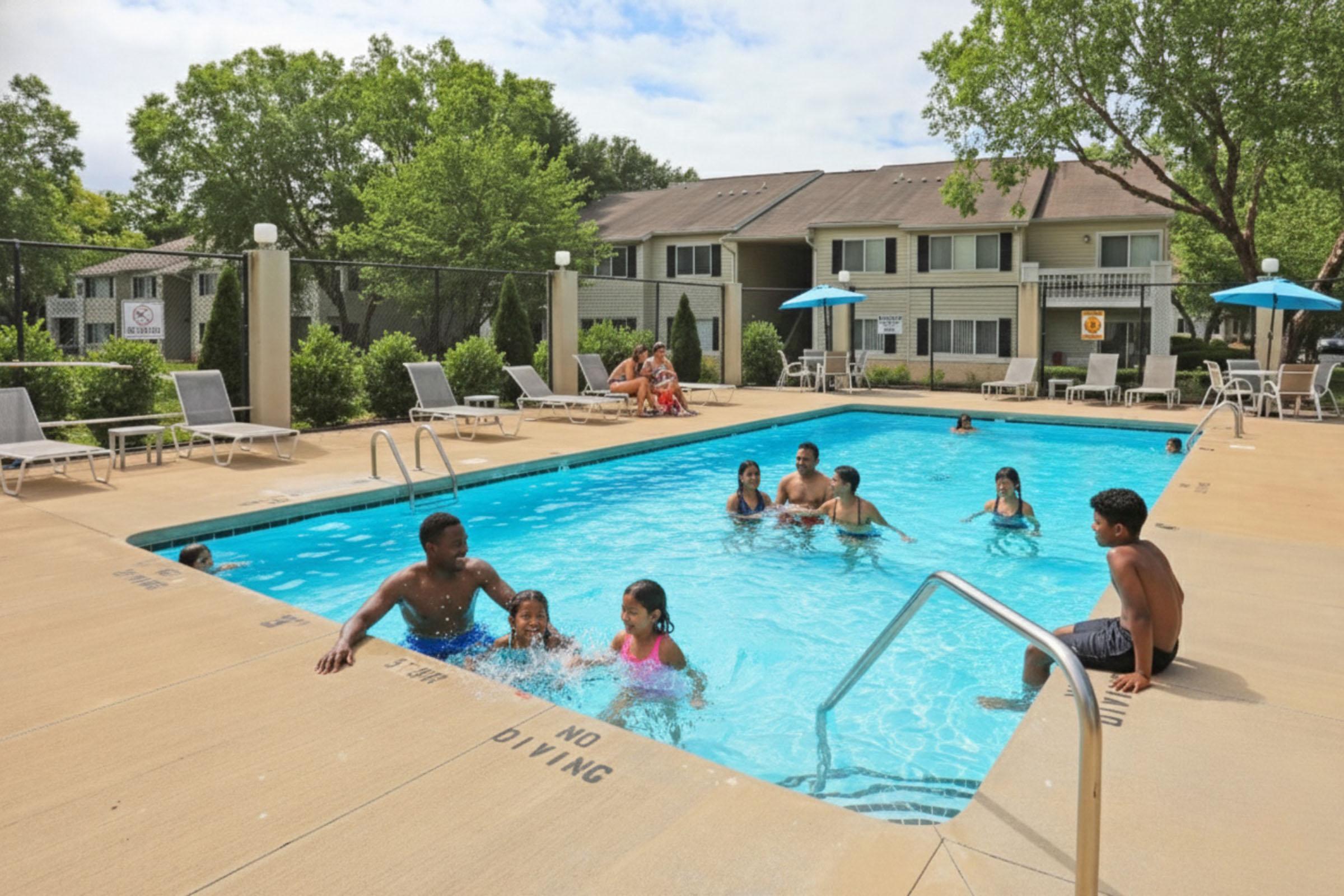 A sunny day at a community pool with several people enjoying the water. Some children are swimming and playing, while adults relax on lounge chairs nearby. Umbrellas provide shade, and residential buildings can be seen in the background. The scene conveys a lively, family-friendly atmosphere.