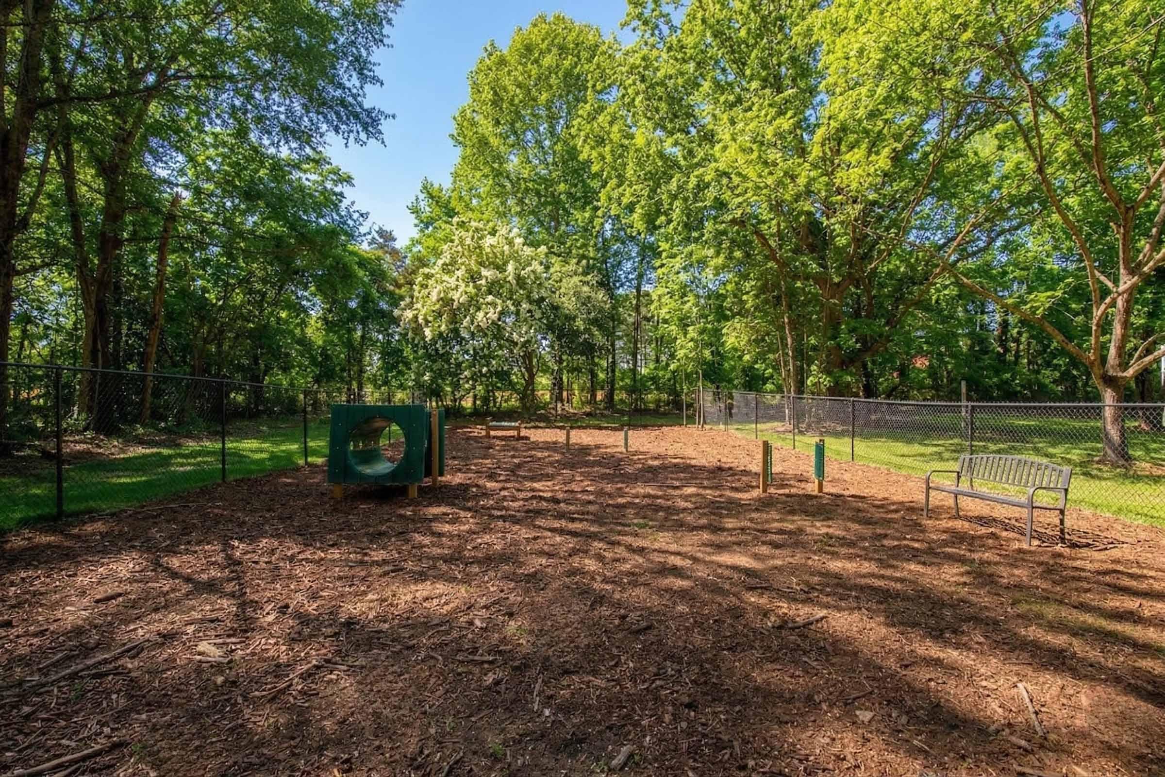 A sunny dog park with a fenced area featuring a small green dog play structure, scattered mulch on the ground, and a solitary bench. Surrounding trees provide shade, with green foliage and a few flowering plants visible in the background. The overall atmosphere is serene and spacious, ideal for pets to play.