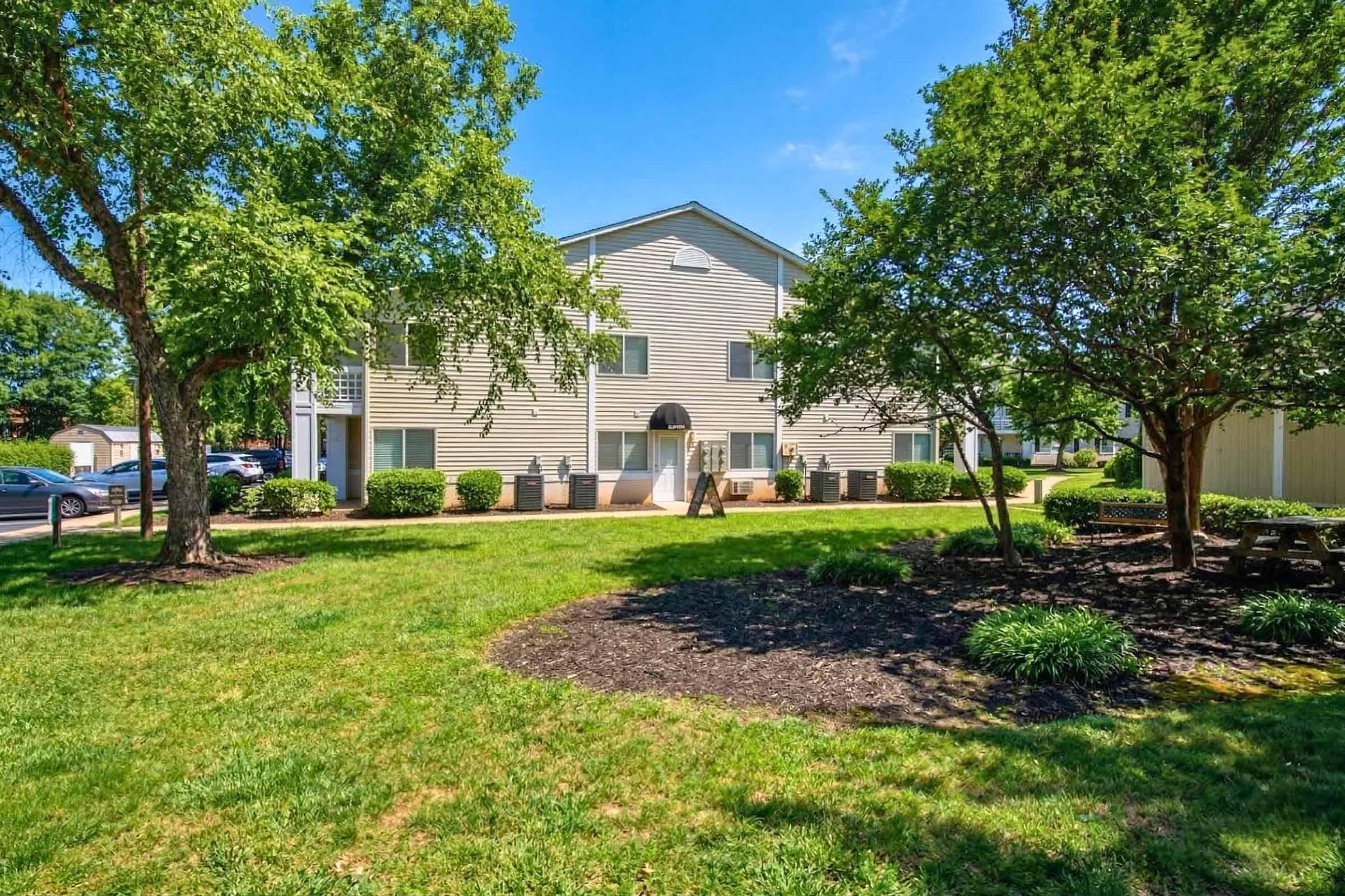 A well-maintained residential building surrounded by lush green trees and landscaped grass. The exterior features light-colored siding, and there are parking spaces visible in the background. The scene is bright and sunny, highlighting the inviting natural environment.
