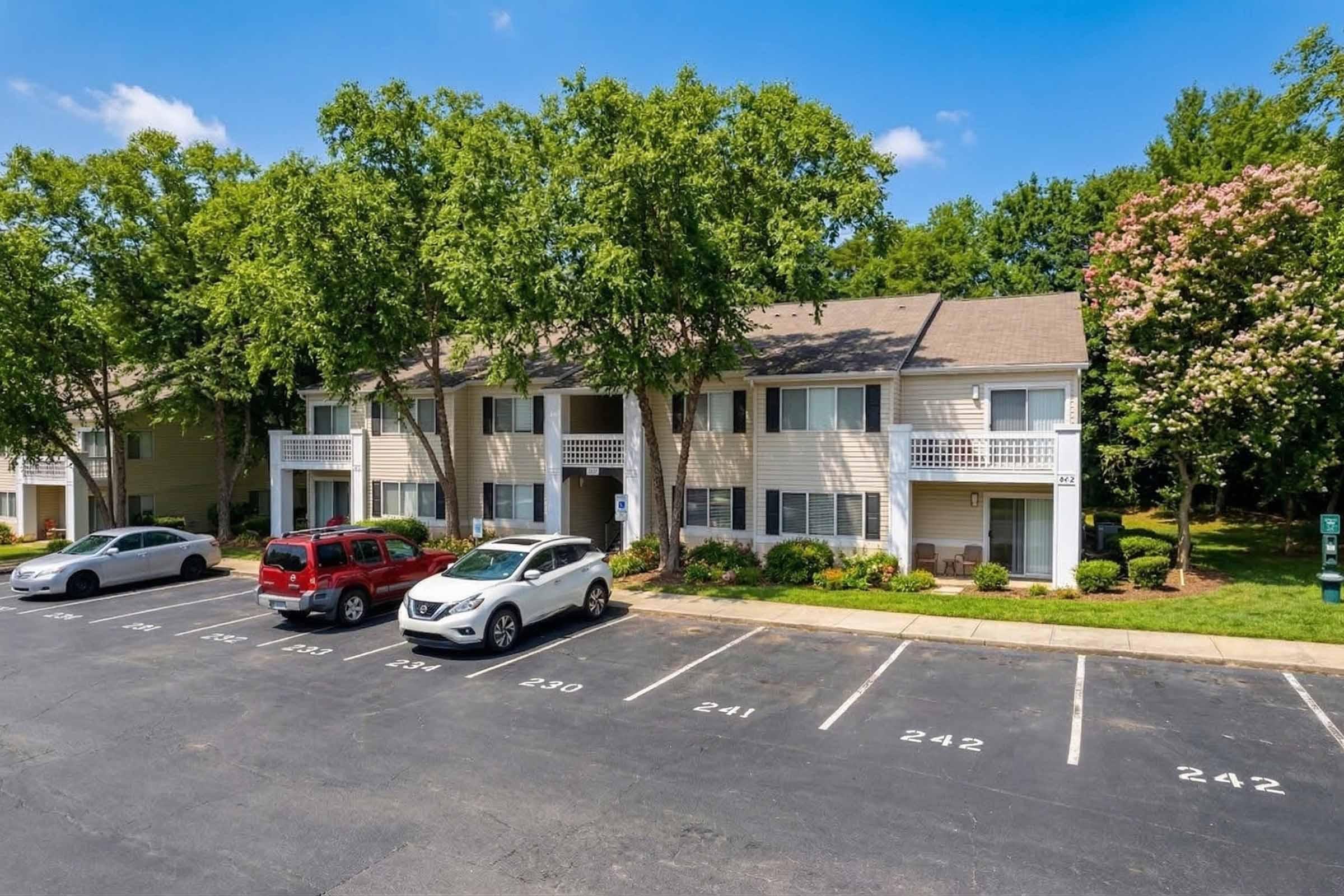 A two-story apartment building surrounded by trees and greenery. The facade features multiple windows and balconies. Several parked cars are visible in the foreground, with a clear blue sky in the background, suggesting a sunny day.