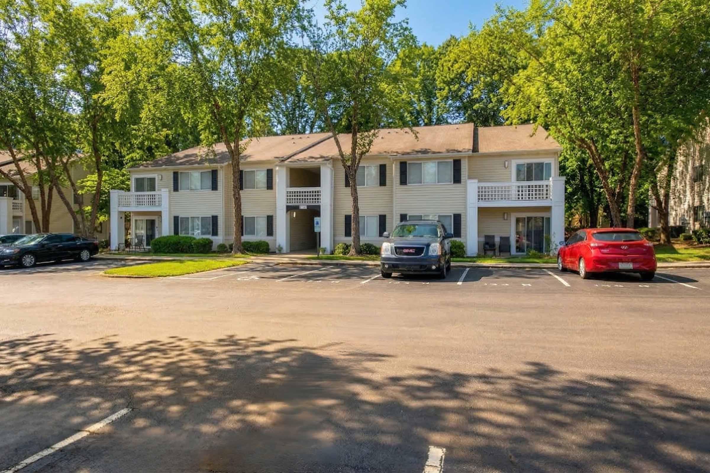 Two-story apartment building surrounded by trees, with several parking spaces in front. The building features a light-colored facade with balconies. Two cars are parked in the lot, one silver and one red, under a sunny sky.