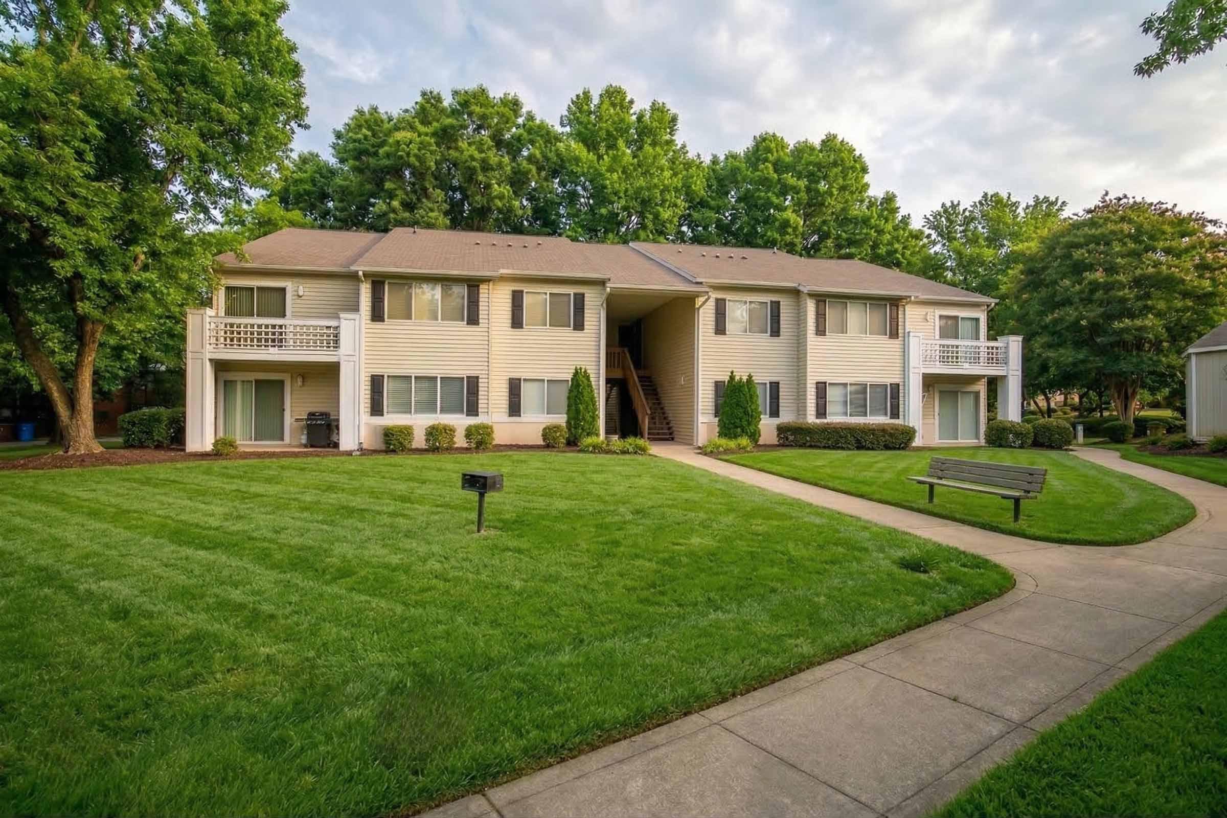 Two-story apartment building surrounded by well-maintained lawns and trees. The building features a light-colored exterior with balconies on the upper level. A pathway runs alongside the building, leading to a bench and neatly trimmed hedges. The area is lush and green, suggesting a peaceful residential setting.