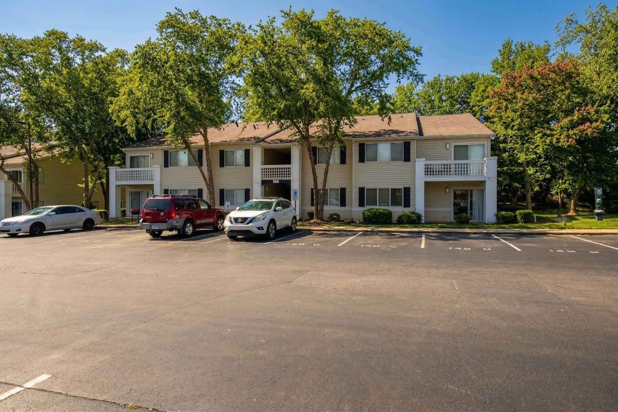 A row of two-story apartment buildings surrounded by greenery. The parking lot in front has several vehicles, including a red SUV and a white car, with trees providing shade in the background. The buildings feature balconies and are set against a clear blue sky.
