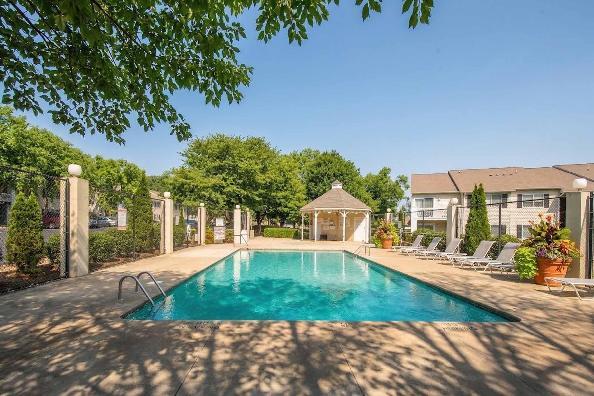 A clear swimming pool surrounded by lounge chairs and a gazebo. Lush greenery and trees are visible in the background, creating a serene outdoor environment. The sun is shining, indicating a bright day. Ideal for relaxation and leisure activities.