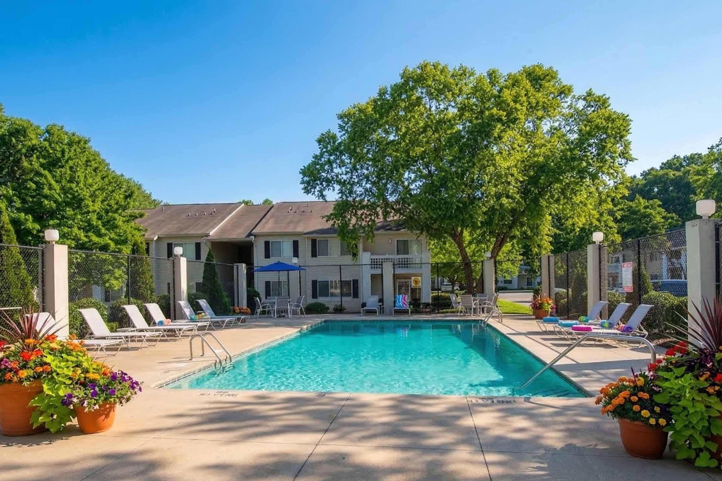 A clear blue swimming pool surrounded by lounge chairs, potted plants, and a large tree. In the background, a multi-unit residential building can be seen, along with vibrant landscaping and a fenced area. The scene is sunny and inviting, perfect for relaxation.