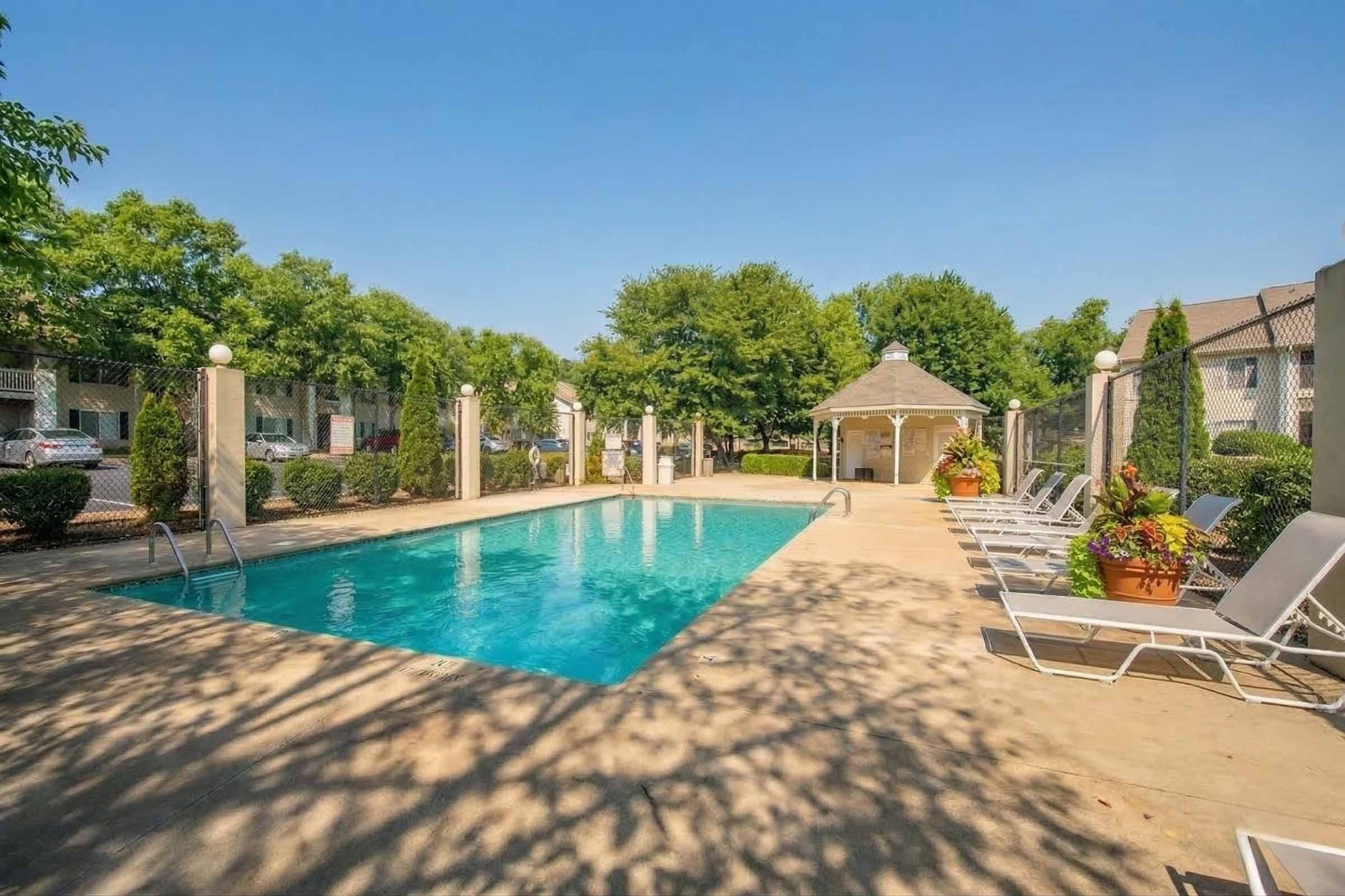 A clean swimming pool surrounded by lounge chairs, with a gazebo and lush greenery in the background. The area is fenced, providing a private and inviting atmosphere for relaxation. Bright blue skies enhance the tranquil setting.
