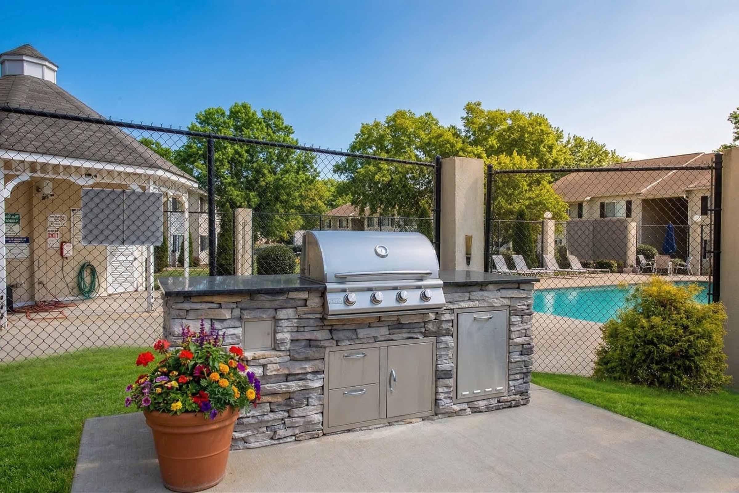 An outdoor grilling station with a stainless steel grill and stone countertop, surrounded by a well-maintained lawn and colorful flower pot. In the background, there is a fenced pool area with lounge chairs and a gazebo, creating a relaxing atmosphere for outdoor gatherings.