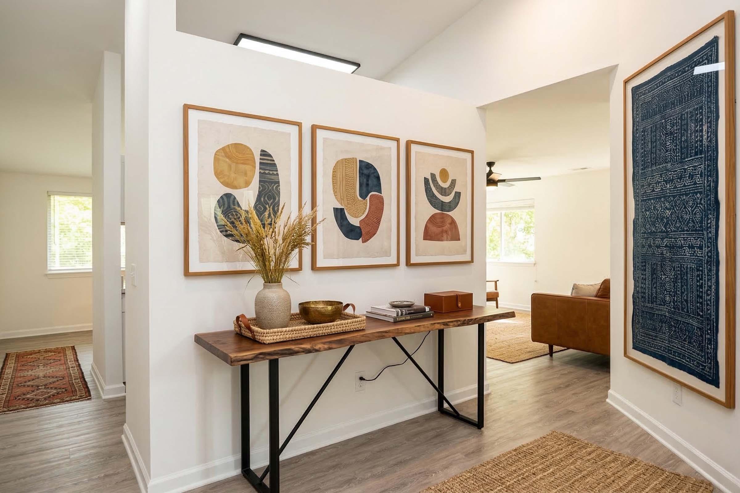 A well-decorated hallway featuring three framed abstract prints on a white wall, a wooden console table with decorative objects like a vase of dried flowers and a bowl. A rug is visible on the floor, and a glimpse of a cozy living room with a brown chair can be seen in the background.