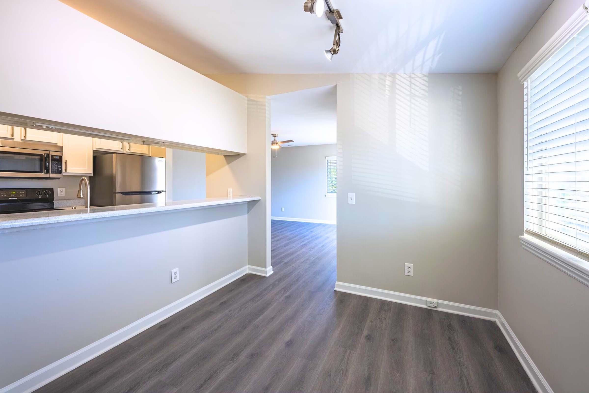 Bright and spacious interior of an apartment, featuring an open kitchen with a bar area, sleek countertops, and stainless steel appliances. The living area is visible through a doorway, showcasing large windows that provide natural light. Light-colored walls and modern flooring create a welcoming atmosphere.