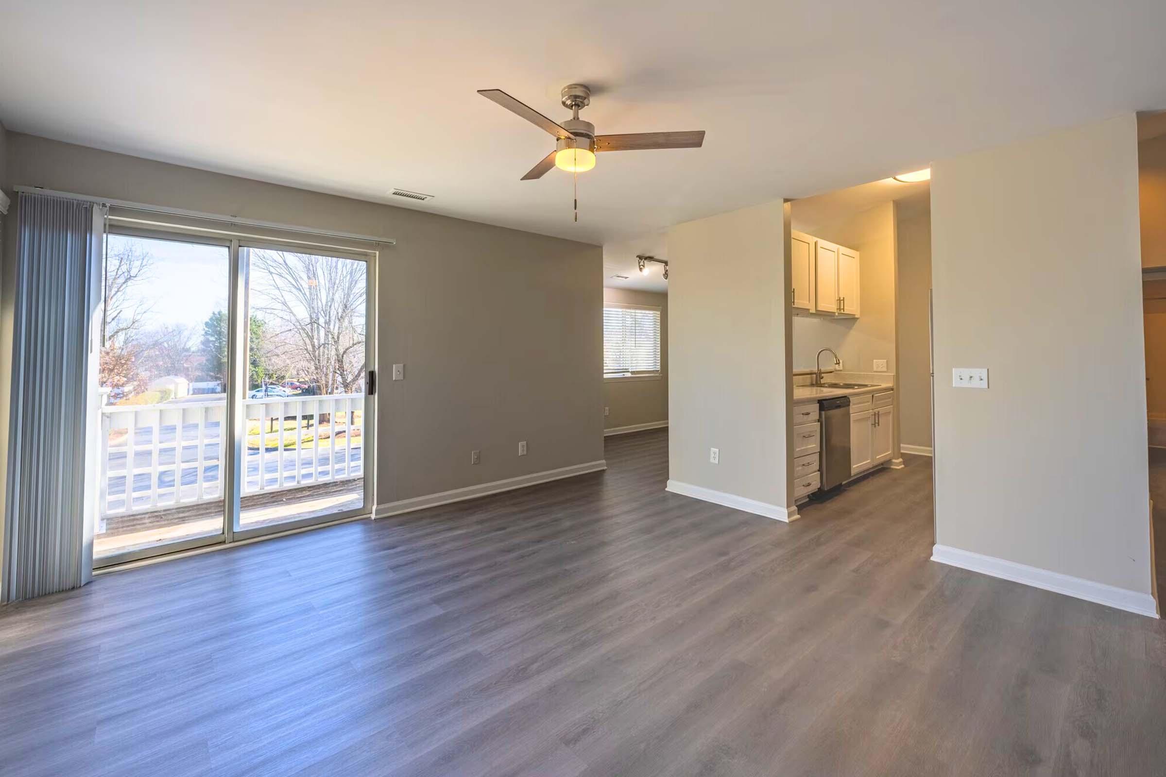 Spacious living area featuring a ceiling fan, large windows for natural light, and an open kitchen space. Sliding glass doors lead to a balcony, and the room has neutral-colored walls and modern flooring.