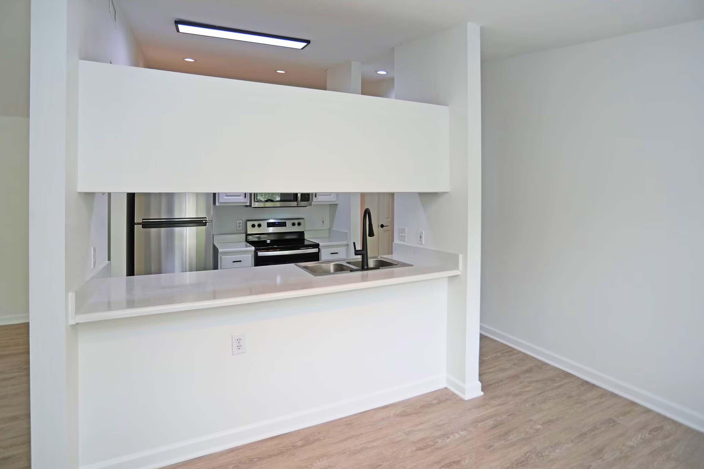 Modern kitchen with a clean, minimalistic design. Features a white countertop, stainless steel sink, and a window to the living area. The kitchen has a stainless steel stove and fridge, with bright lighting and light-colored walls, enhancing the spacious feel of the open layout.