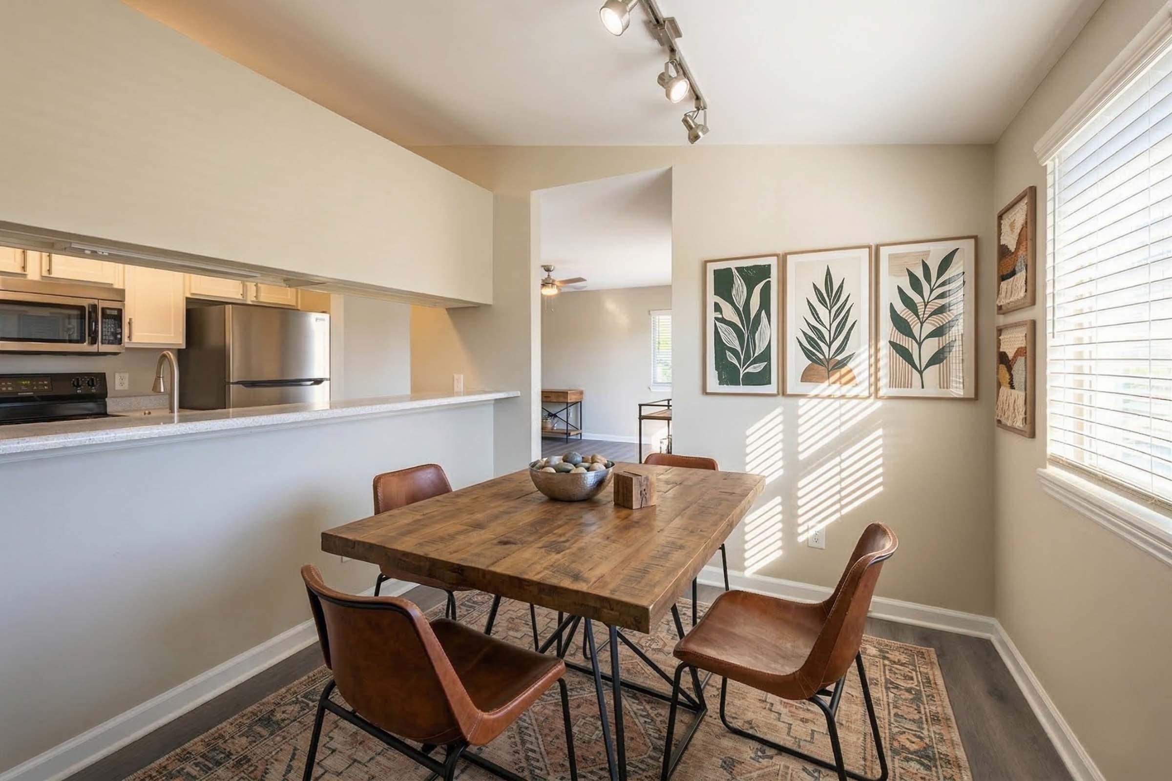 A cozy dining area featuring a wooden table surrounded by four leather chairs. A bowl in the center of the table adds a decorative touch. The walls are adorned with three framed botanical prints. Natural light streams in through a window, creating a warm and inviting atmosphere.
