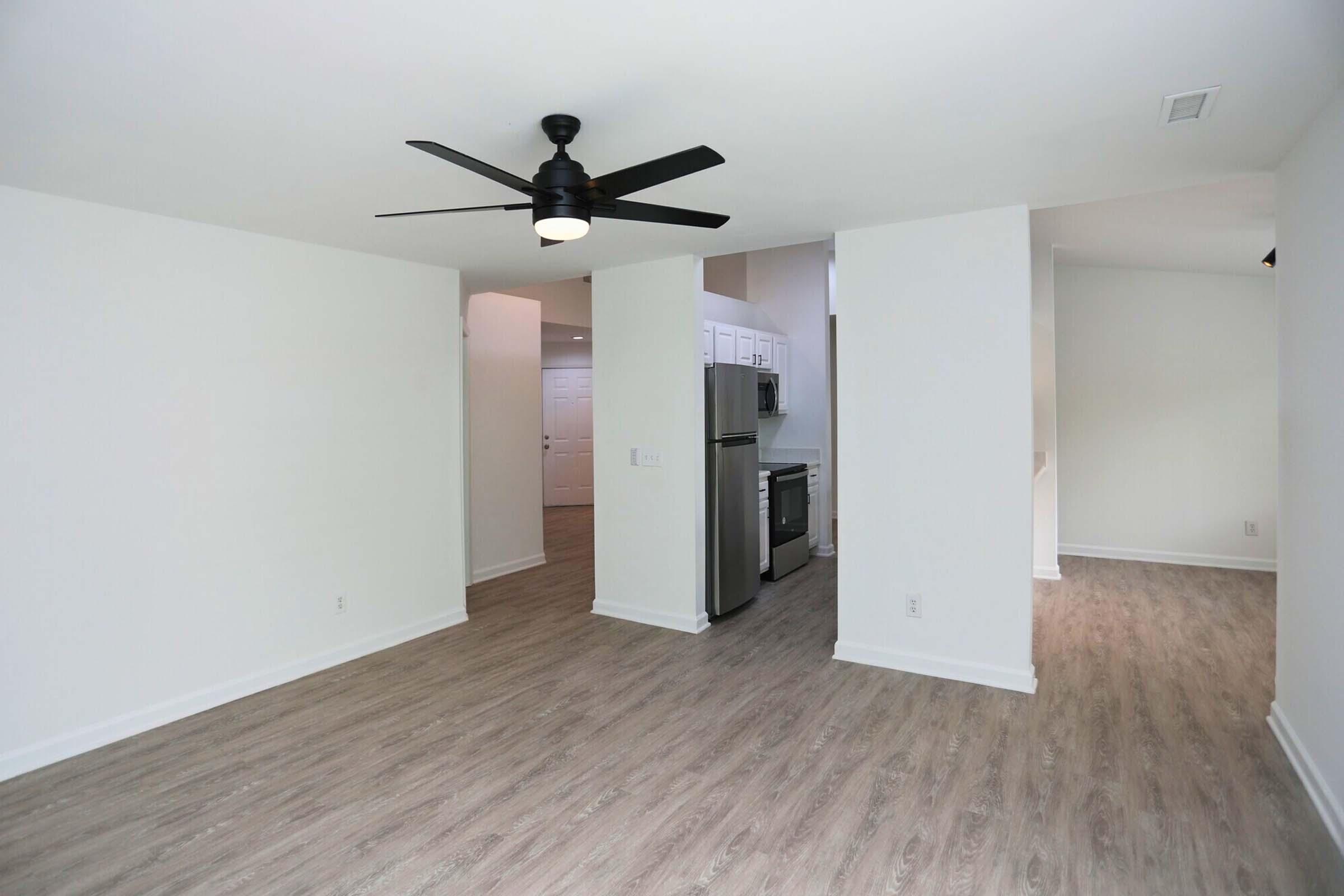 Modern, spacious living area featuring light-colored walls and wood-style flooring. A ceiling fan is mounted in the center, and an open kitchen space is visible in the background, showcasing stainless steel appliances and white cabinetry. The overall aesthetic is bright and minimalist.