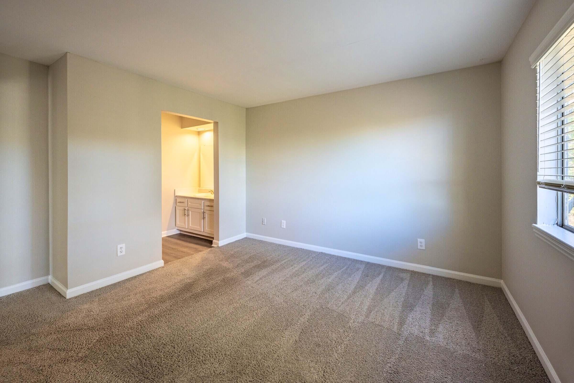 A spacious, empty room with neutral-colored walls and natural light from a window. The flooring is carpeted, and there is an open doorway leading to a bathroom area with light-colored cabinetry. The overall space appears clean and modern, suitable for various furnishing options.