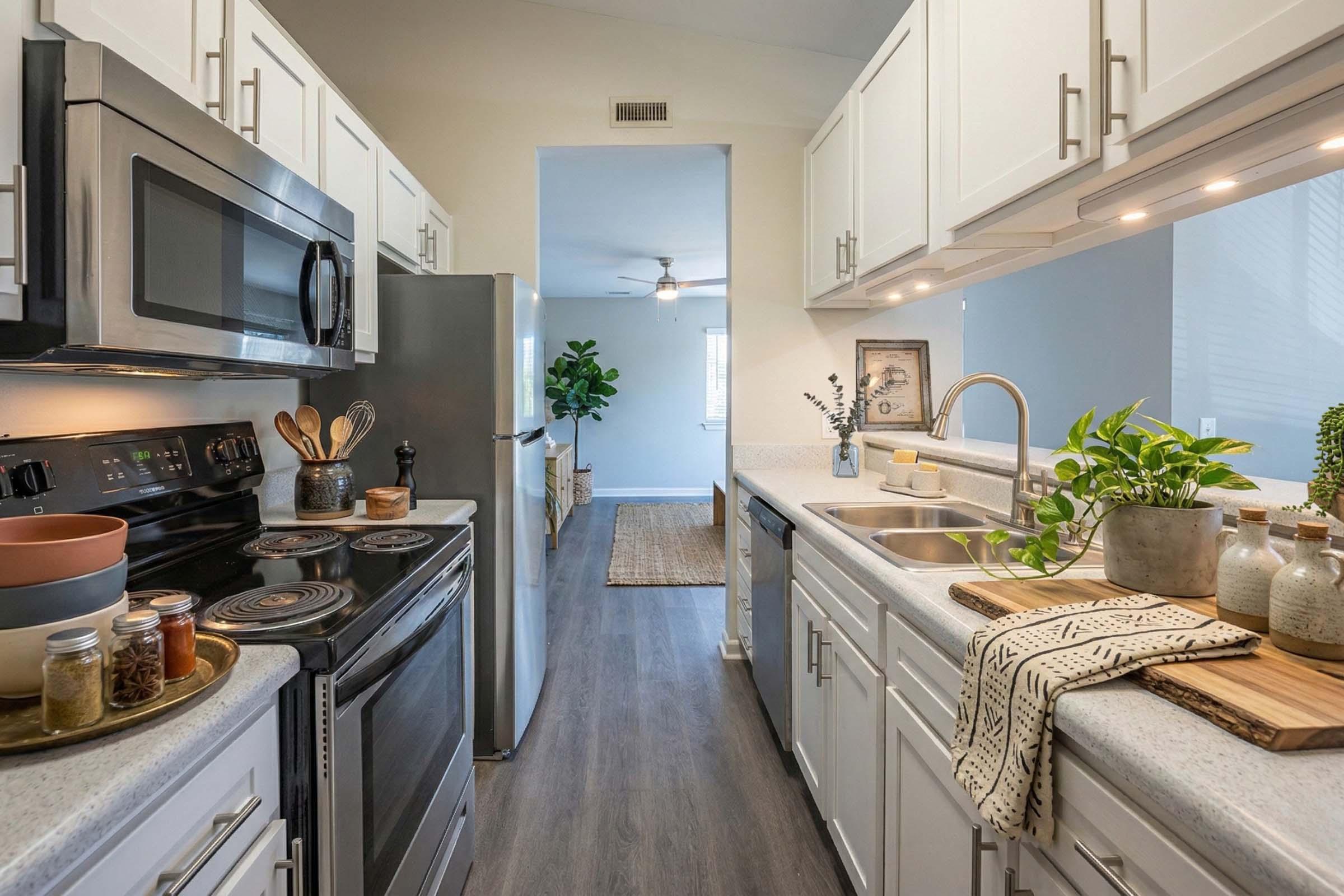 A modern kitchen featuring stainless steel appliances, including a microwave and stove, along with white cabinets and gray countertops. The kitchen is well-lit and includes decorative elements like plants, cutting boards, and jars. A living area is visible through an open door at the far end.