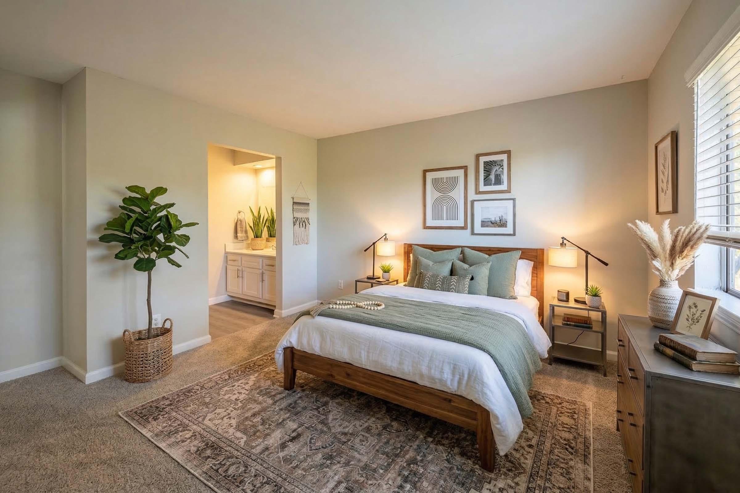 A modern bedroom featuring a wooden bed with cozy bedding and decorative pillows. There are framed artworks on the wall, a small bedside table with lamps, and a dresser. A potted plant adds greenery, and the room has a warm, inviting atmosphere with neutral tones and a stylish area rug. A bathroom entrance is visible in the background.