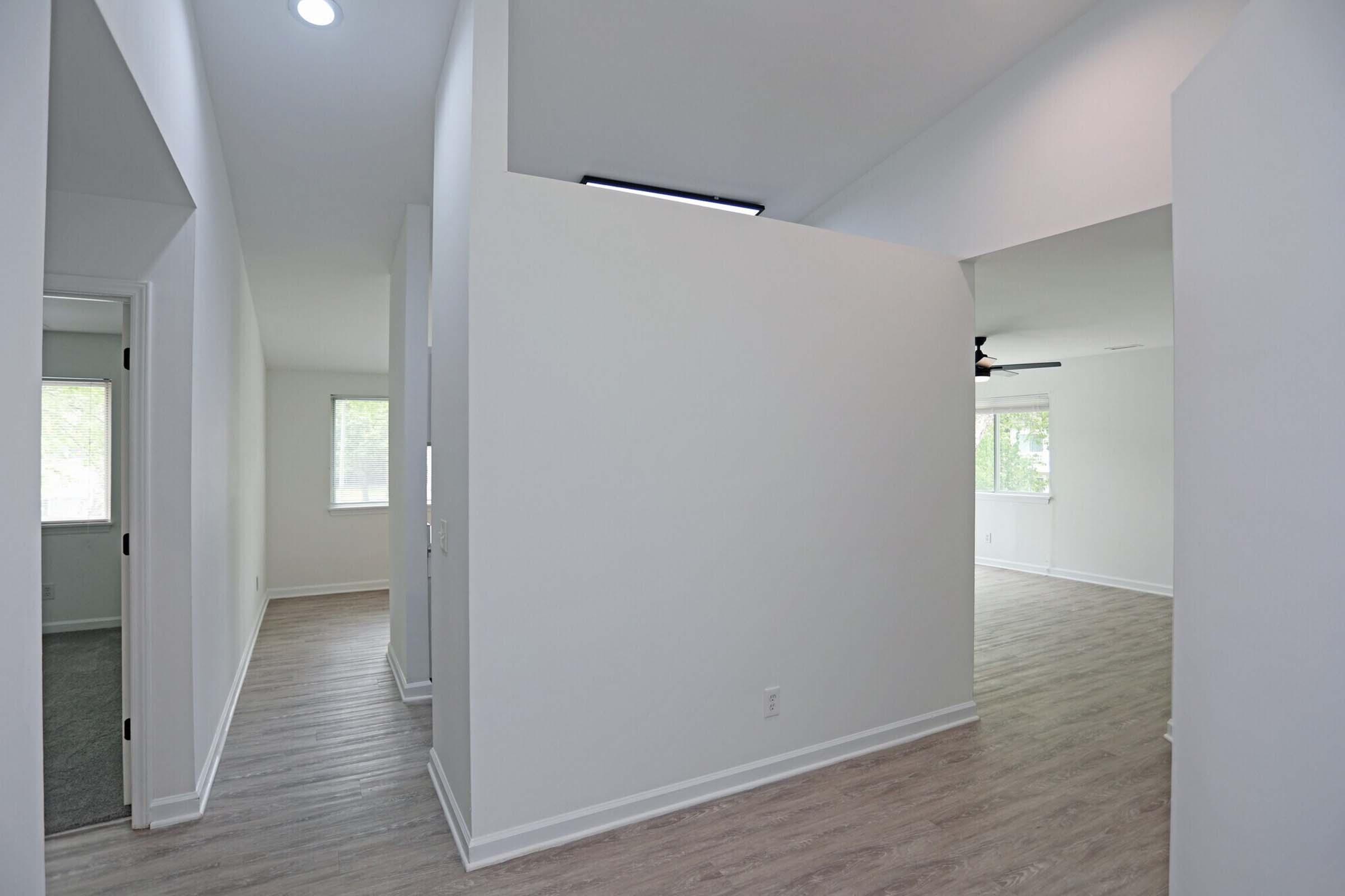 A bright and open interior space featuring light-colored walls, modern recessed lighting, and hardwood flooring. The layout includes a hallway leading to a spacious area with large windows, providing natural light. The scene conveys a fresh, contemporary living environment.