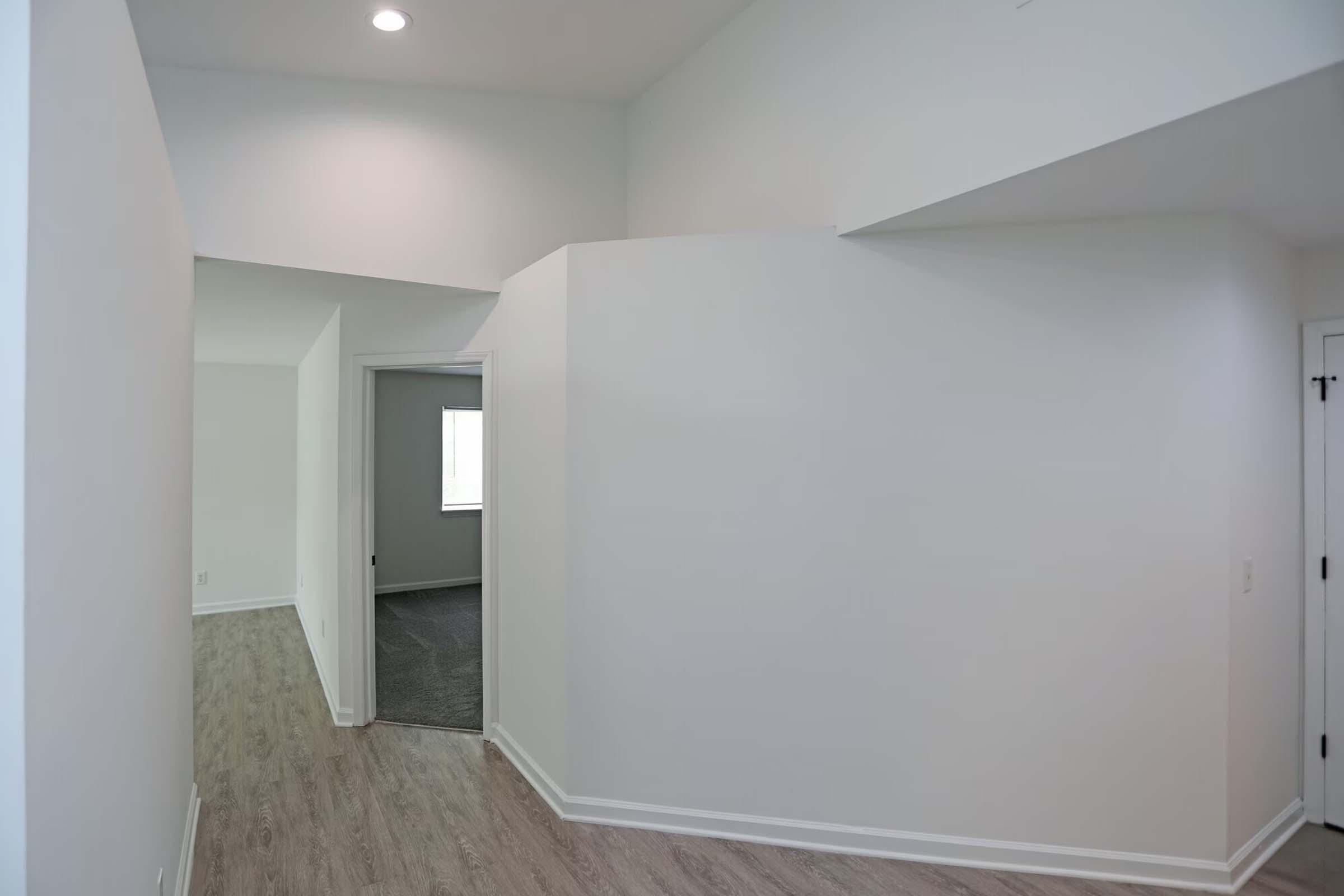 A modern interior space featuring light-colored walls and a light wood floor, with a doorway leading to another room. The area is bright, with natural light coming from a window, and has an open, spacious feel.