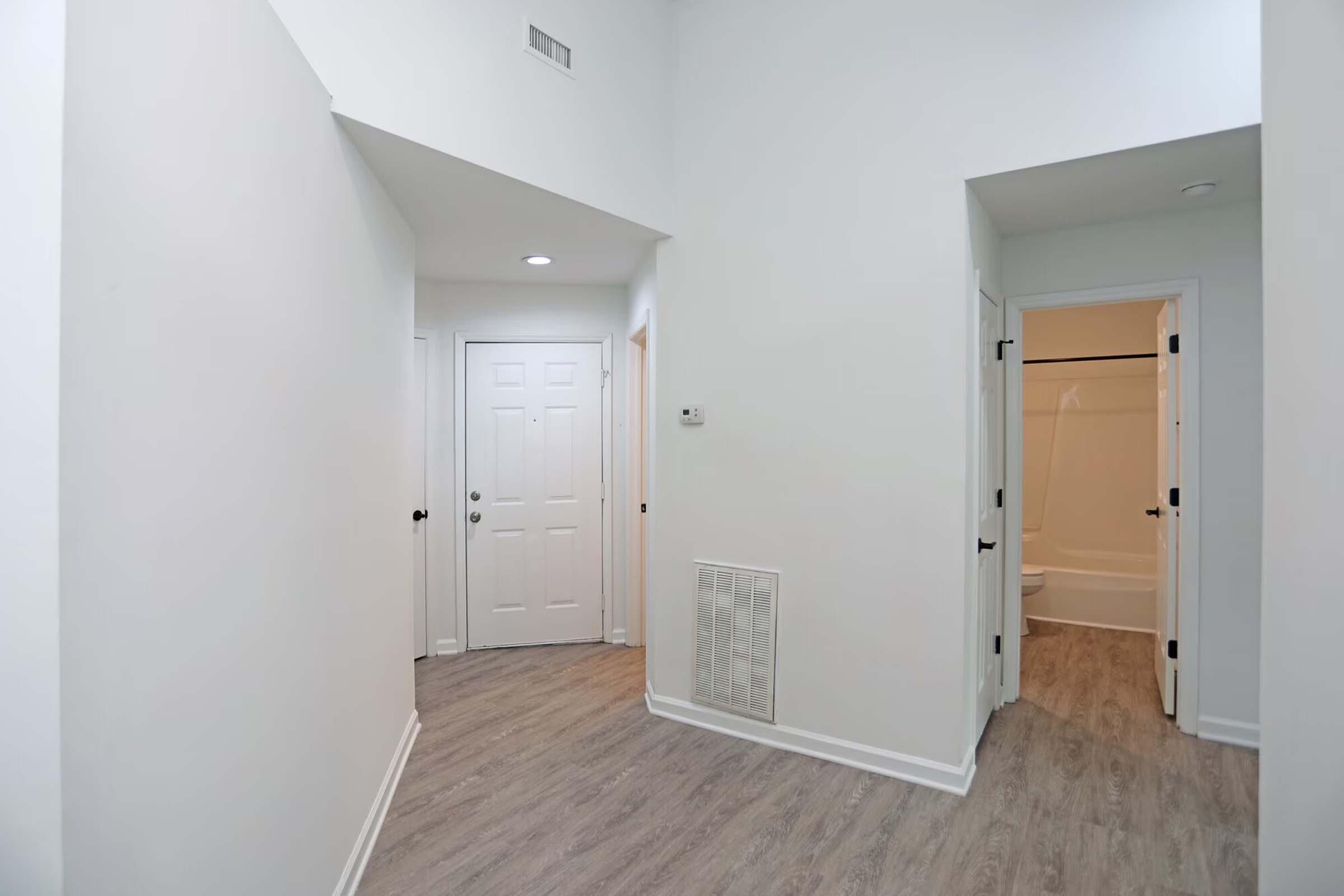 Interior view of a modern hallway leading to a front door and bathrooms. The walls are painted white, and the flooring is light-colored wood laminate. One doorway on the right leads to a bathroom with a bathtub, while the entrance is visible on the left. The space is well-lit and minimalistic.