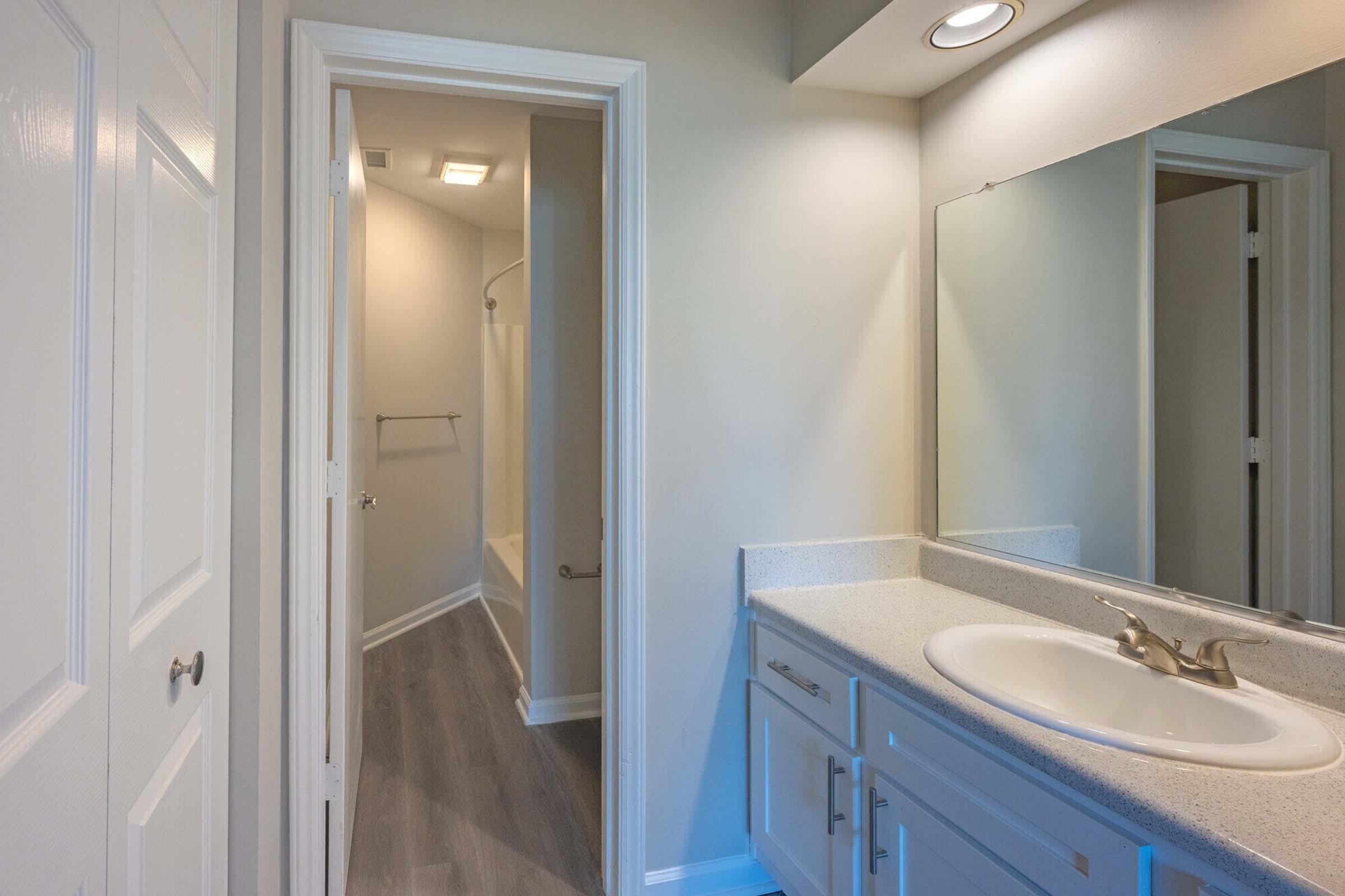 A well-lit bathroom featuring a white countertop with a sink, modern fixtures, and a large mirror. A doorway to a nearby room is visible, leading to a light-colored area with a bathtub. The walls are painted in a neutral tone, and the flooring is a light wood finish.