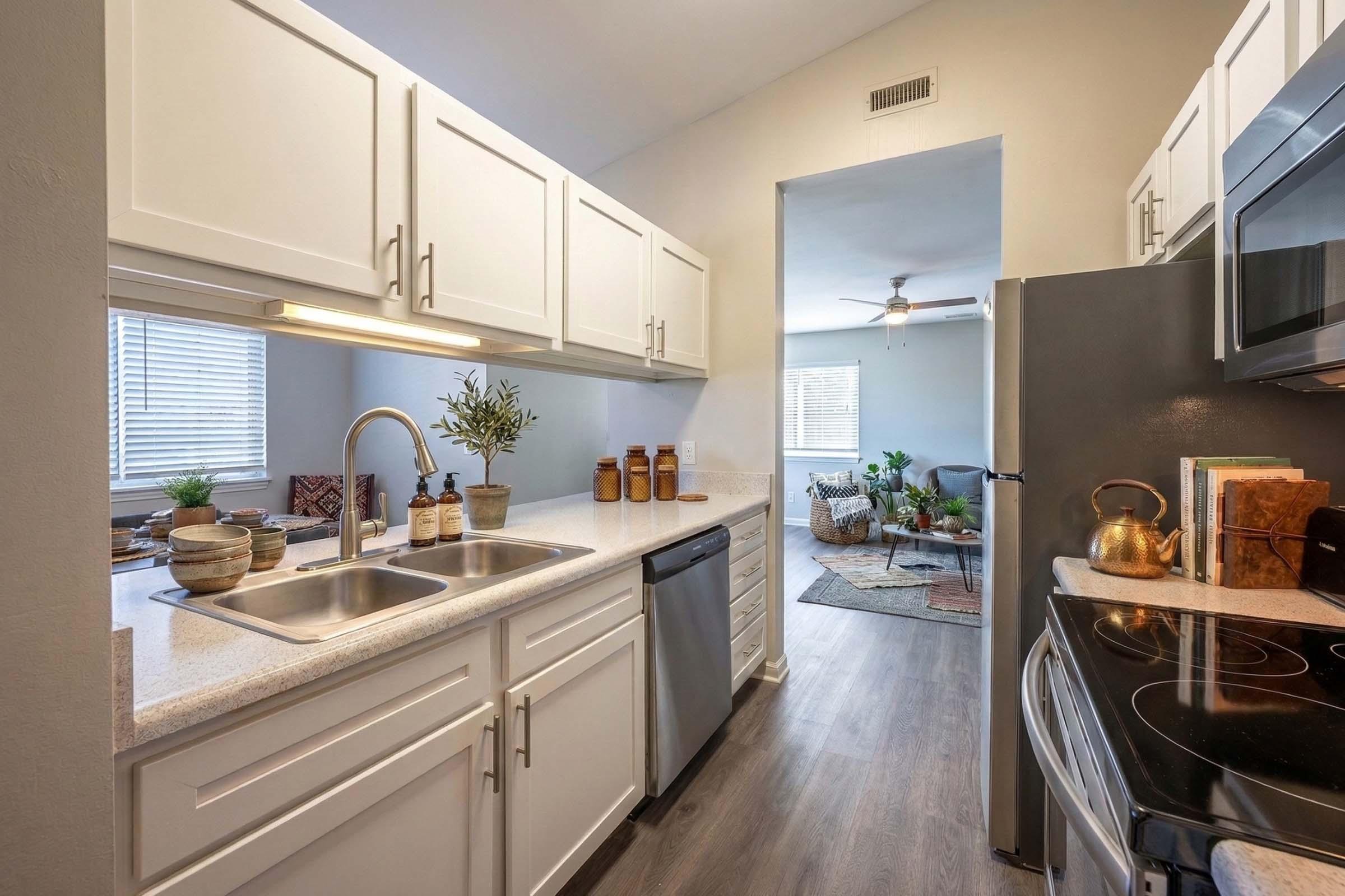 Bright modern kitchen featuring white cabinets, a double sink, a dishwasher, and stainless steel appliances. The countertop is styled with decorative plants and kitchen accessories. View opens to a cozy living area with a fan and comfortable seating, imbued with natural light from nearby windows.