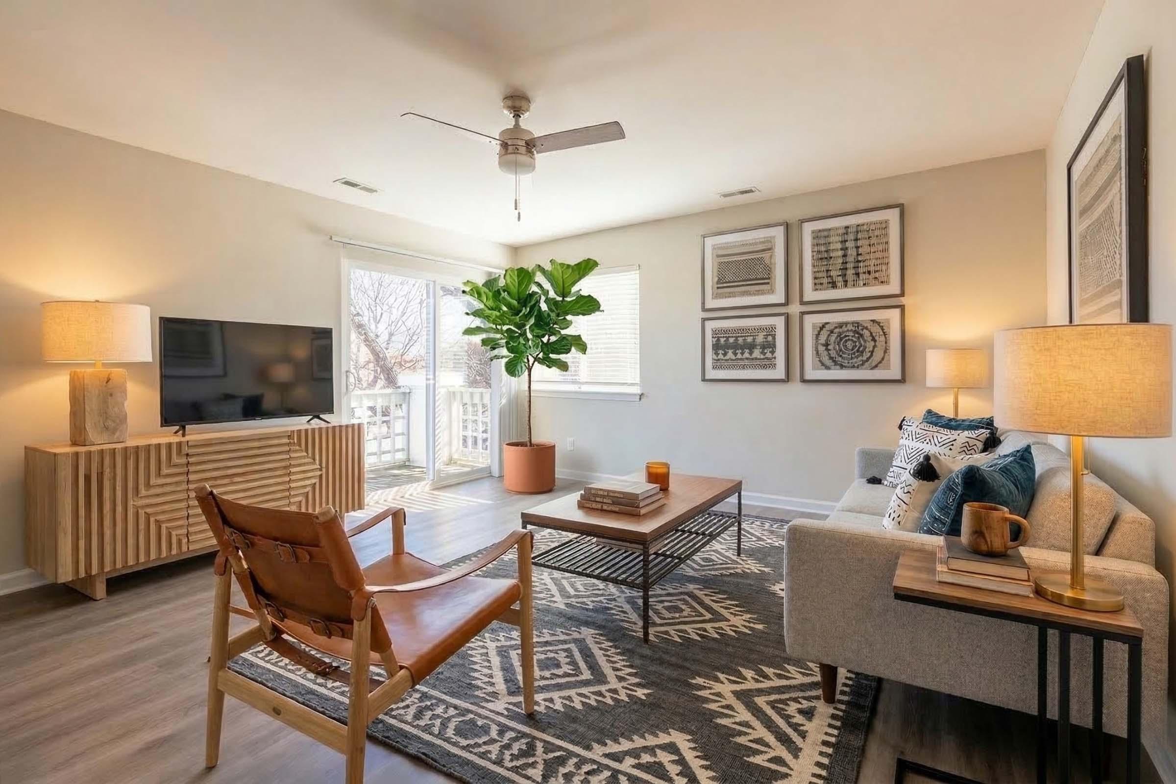 A cozy living room featuring a light gray sofa, a wooden coffee table, and a modern armchair. A large potted plant adds greenery, while wall art and a flat-screen TV provide decor. Soft lighting from two lamps creates a warm ambiance, and a balcony door lets in natural light. A patterned rug completes the space.