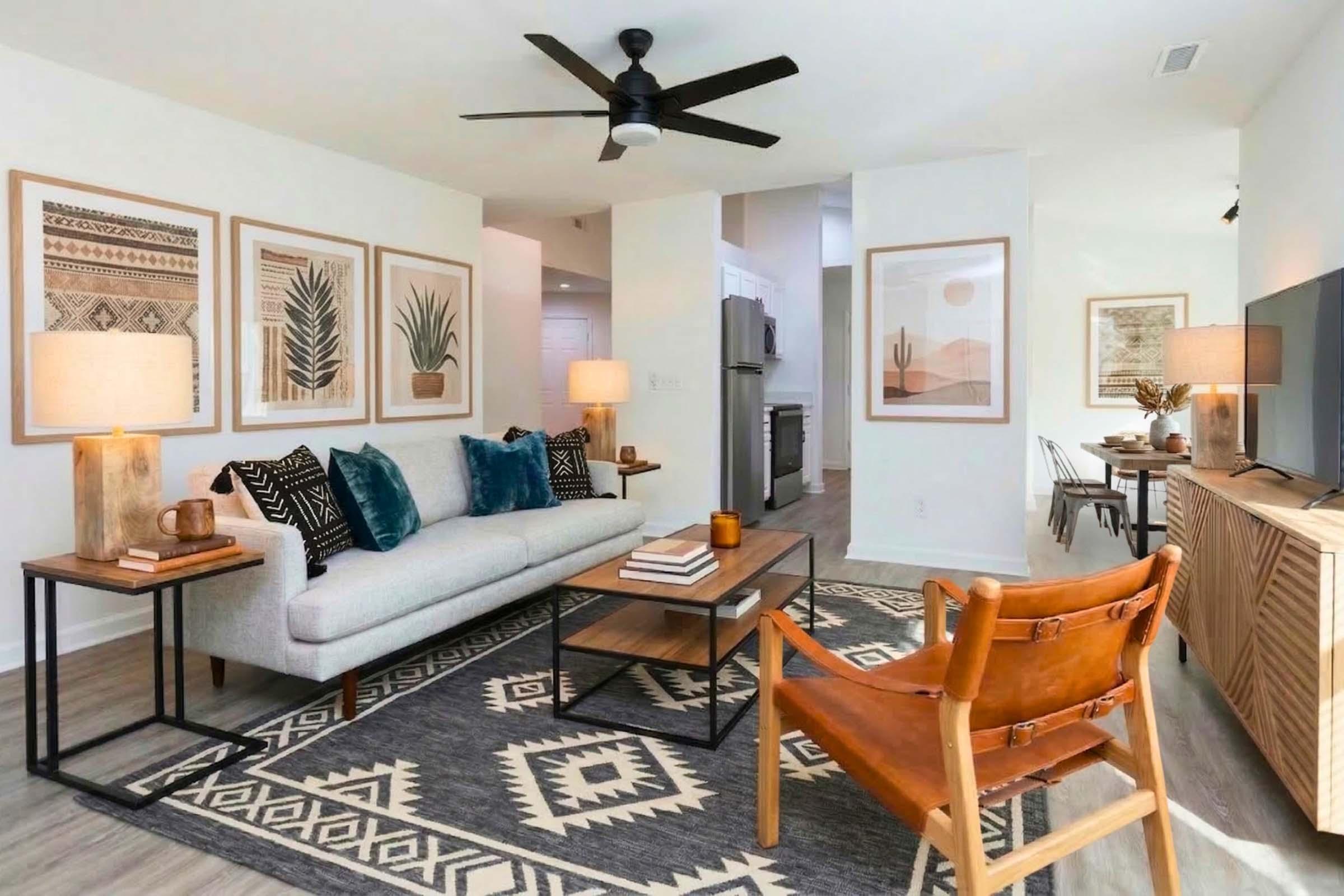 A modern living room featuring neutral tones and stylish furnishings. There is a gray sofa with blue pillows, a wooden coffee table, and a leather chair. The space is decorated with framed art featuring plants and geometric designs, along with lamps on side tables. The floor is adorned with an area rug, and there's a TV console against the wall.