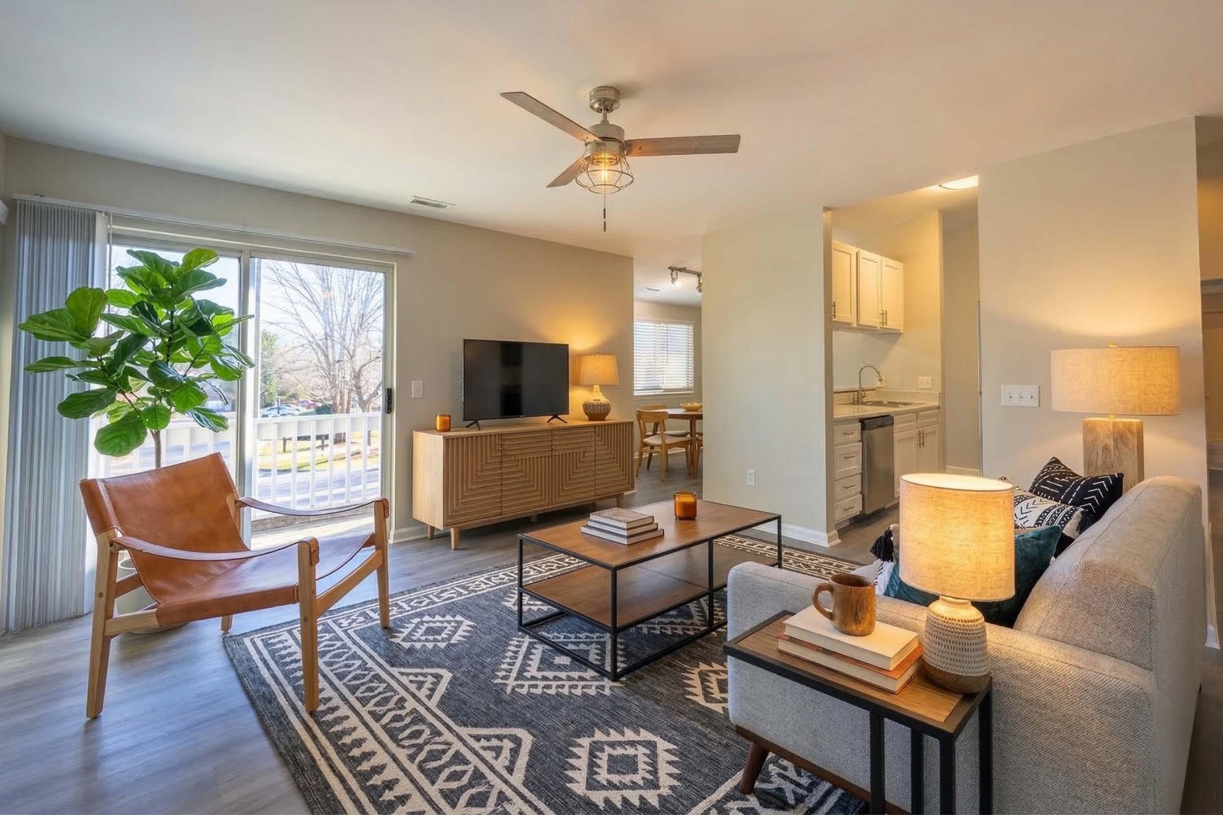 A cozy living room featuring a light-colored sofa, a wooden coffee table, and an accent chair. Large windows let in natural light, and a potted plant adds a touch of greenery. A TV is placed on a media console, with a kitchen visible in the background. The space is decorated with a patterned area rug and warm lighting.