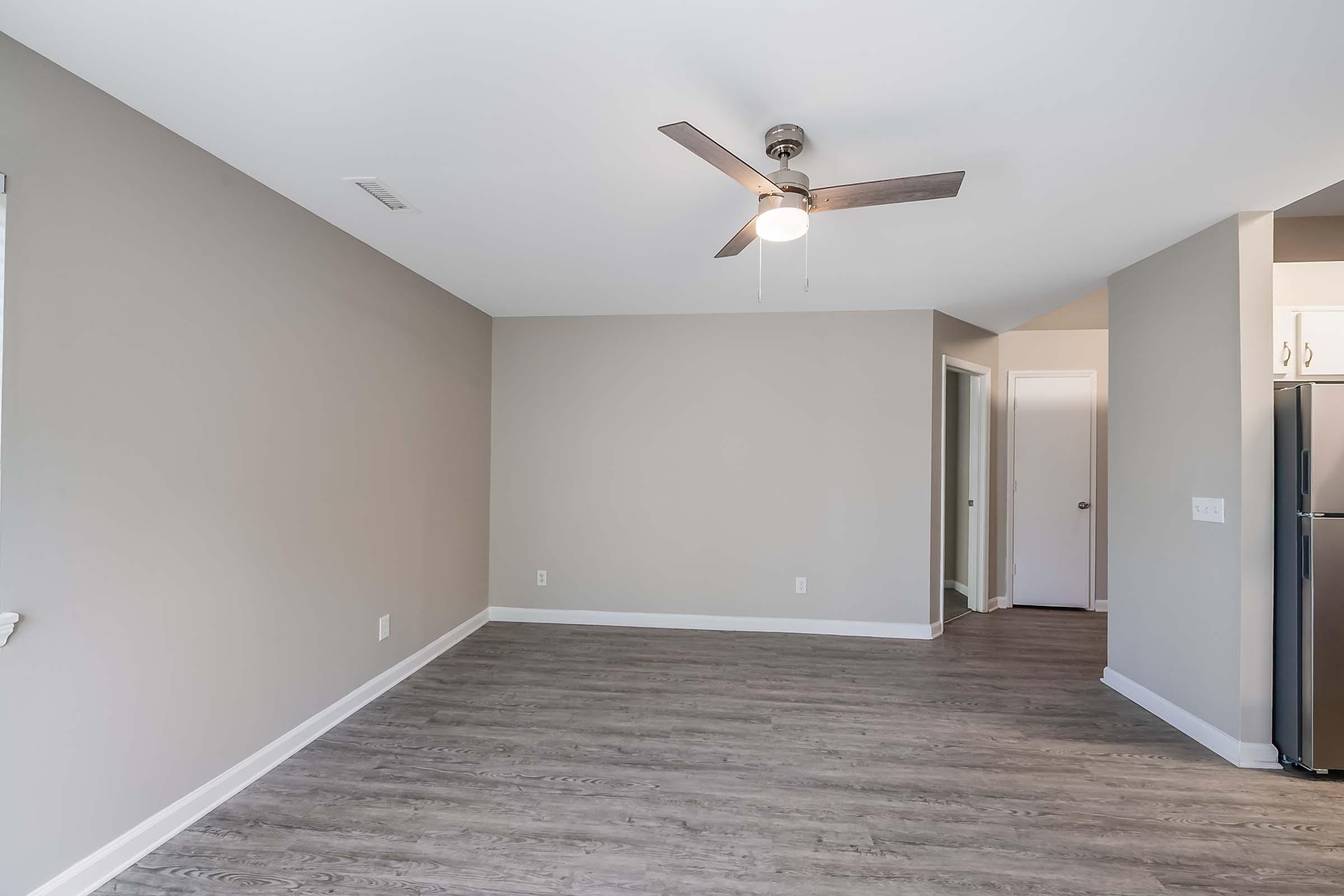 A spacious, light-colored room with a ceiling fan, featuring gray walls and laminate flooring. There is an open layout leading to doorways on the right and left, and a glimpse of a kitchen area with stainless steel appliances in the background. Ideal for a modern living space.