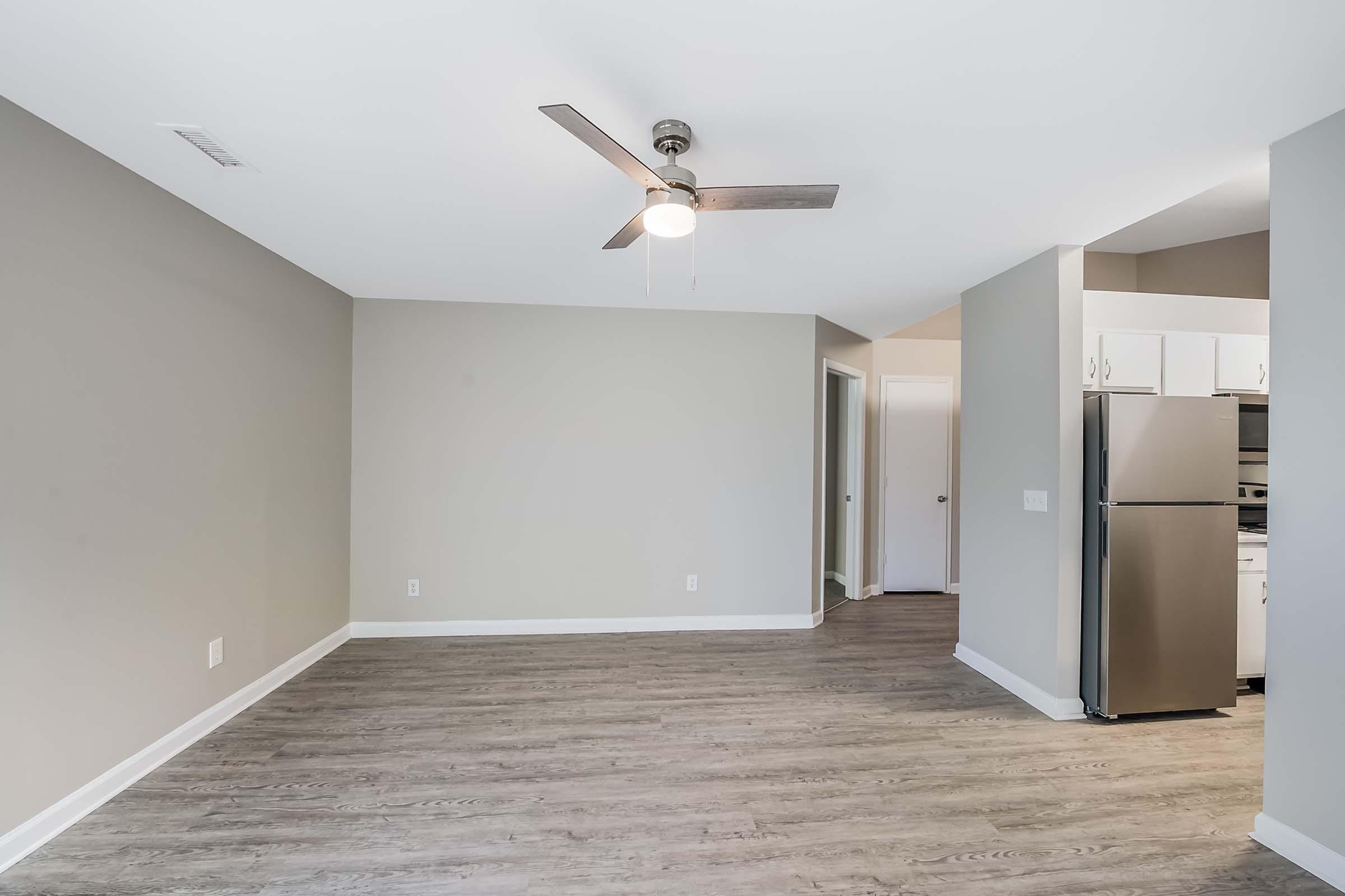 Empty living space featuring a light-colored wall and wood-like flooring. A ceiling fan is installed in the center of the room. To the right, a modern kitchen with white cabinets and stainless-steel appliances is visible, along with a doorway leading to another room.