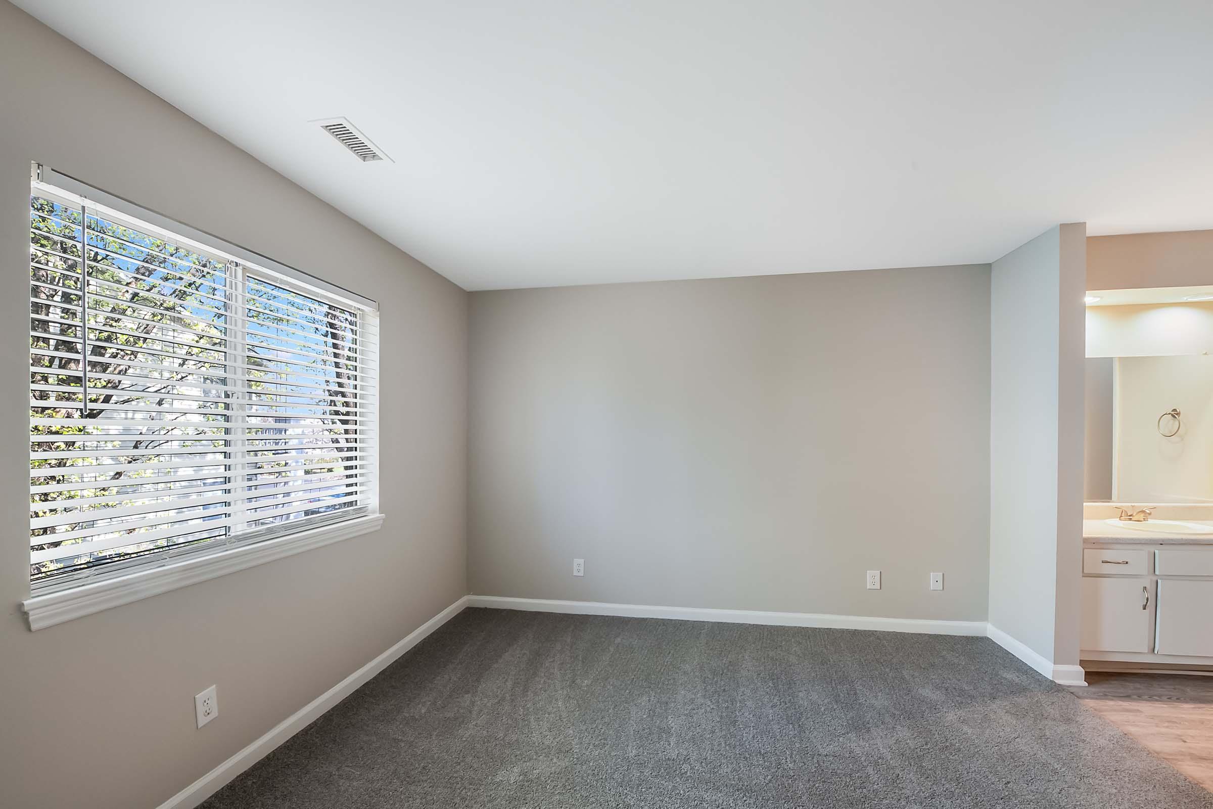 A bright, empty room with light gray walls and a large window featuring white blinds. The floor is covered with soft carpet, and a small area is visible that leads to a bathroom with light-colored fixtures. The overall ambiance is clean and inviting, perfect for potential decoration or furnishing.