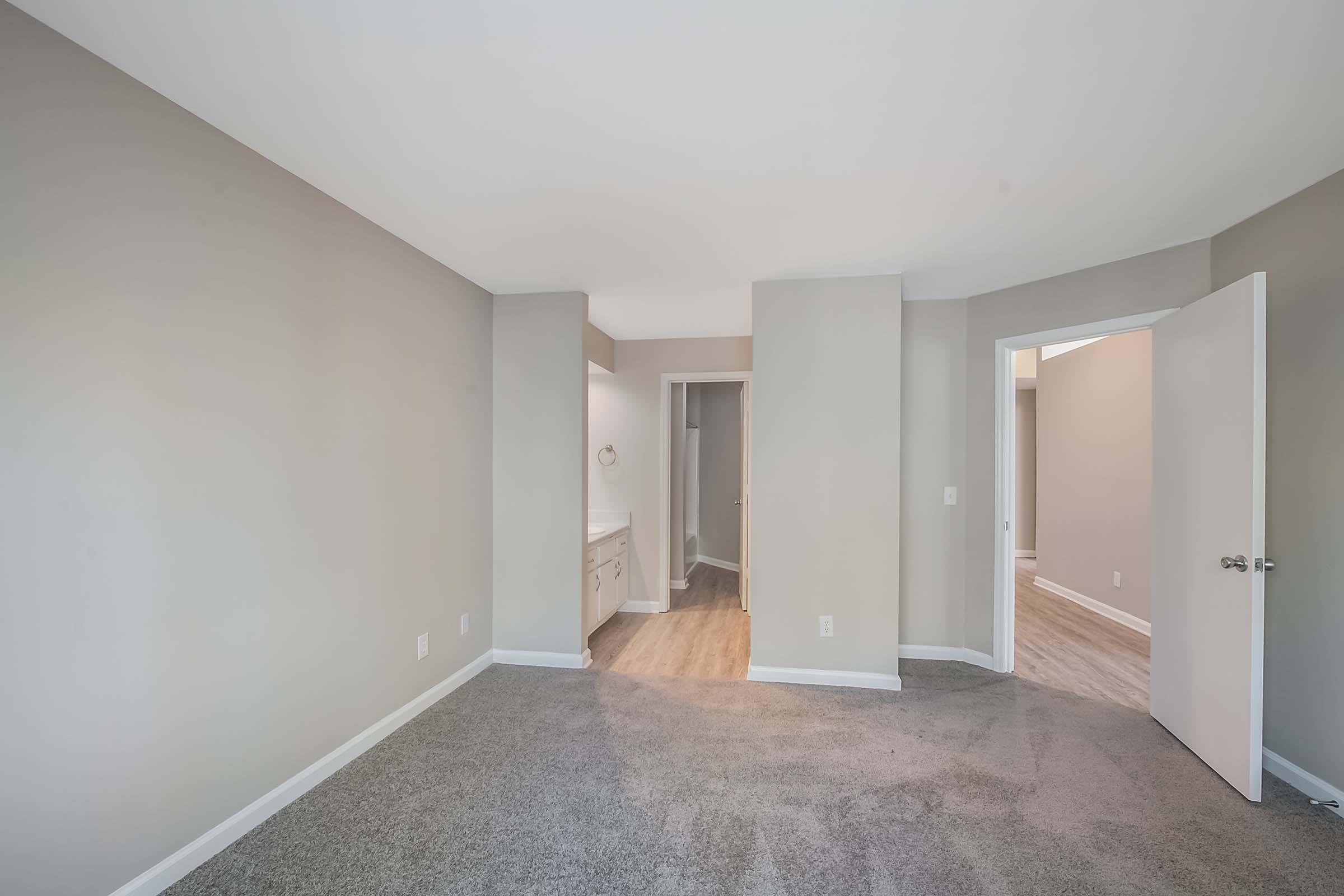 A spacious, empty room with light gray walls and carpeted flooring. Two doorways lead to other areas, and there is a small bathroom vanity visible on the left. The natural light brightens the space, creating an inviting atmosphere.