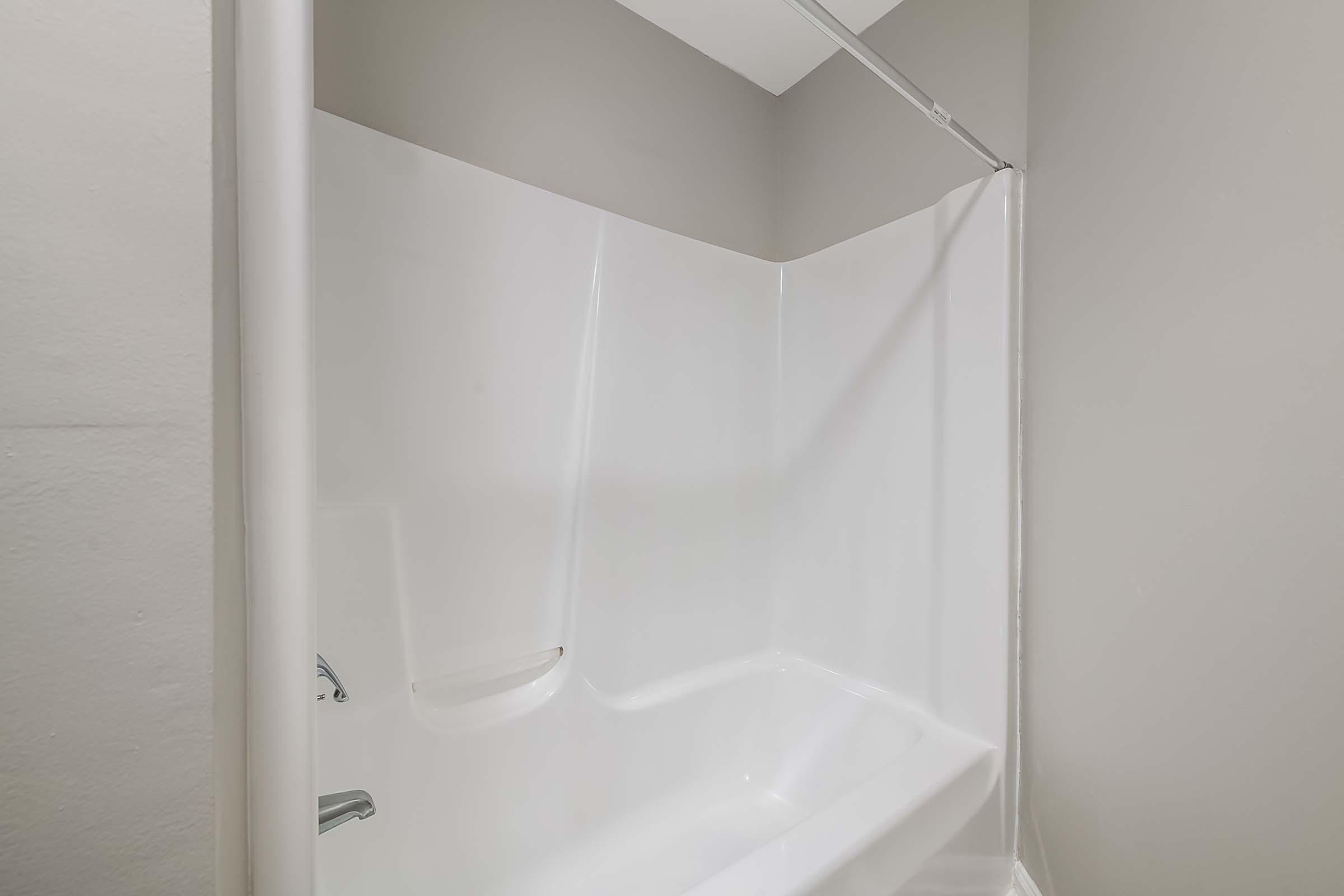 A clean, empty bathtub and shower area with a smooth white surface, set against a light gray wall. The design is modern and simple, featuring a shower curtain rod and no visible fixtures or decorations. The overall atmosphere is minimalistic and uncluttered.