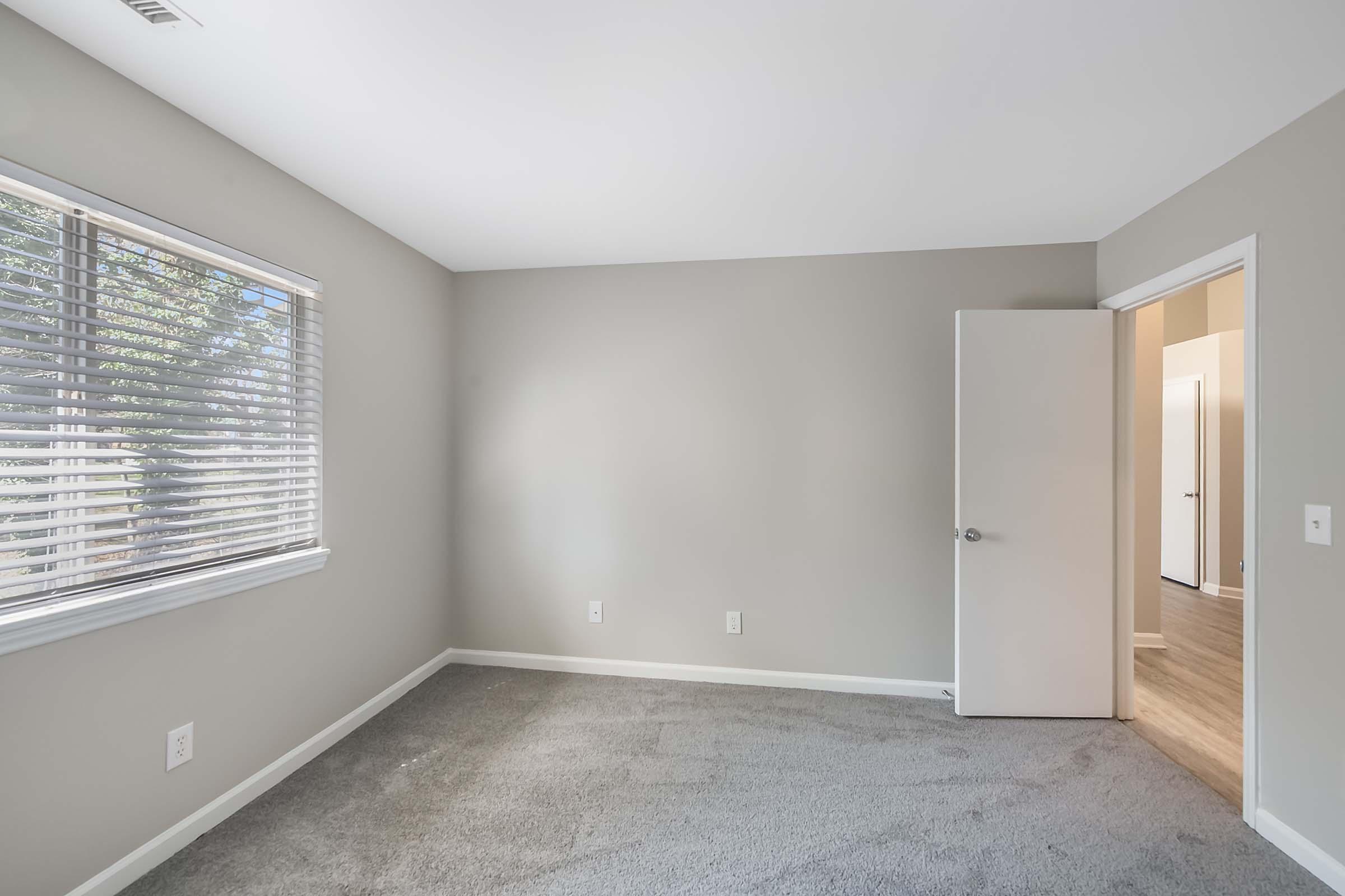A vacant room with light gray walls and a carpeted floor. A window with blinds allows natural light to enter. An open door in the background leads to another room. The space appears clean and minimalist, suitable for various uses.