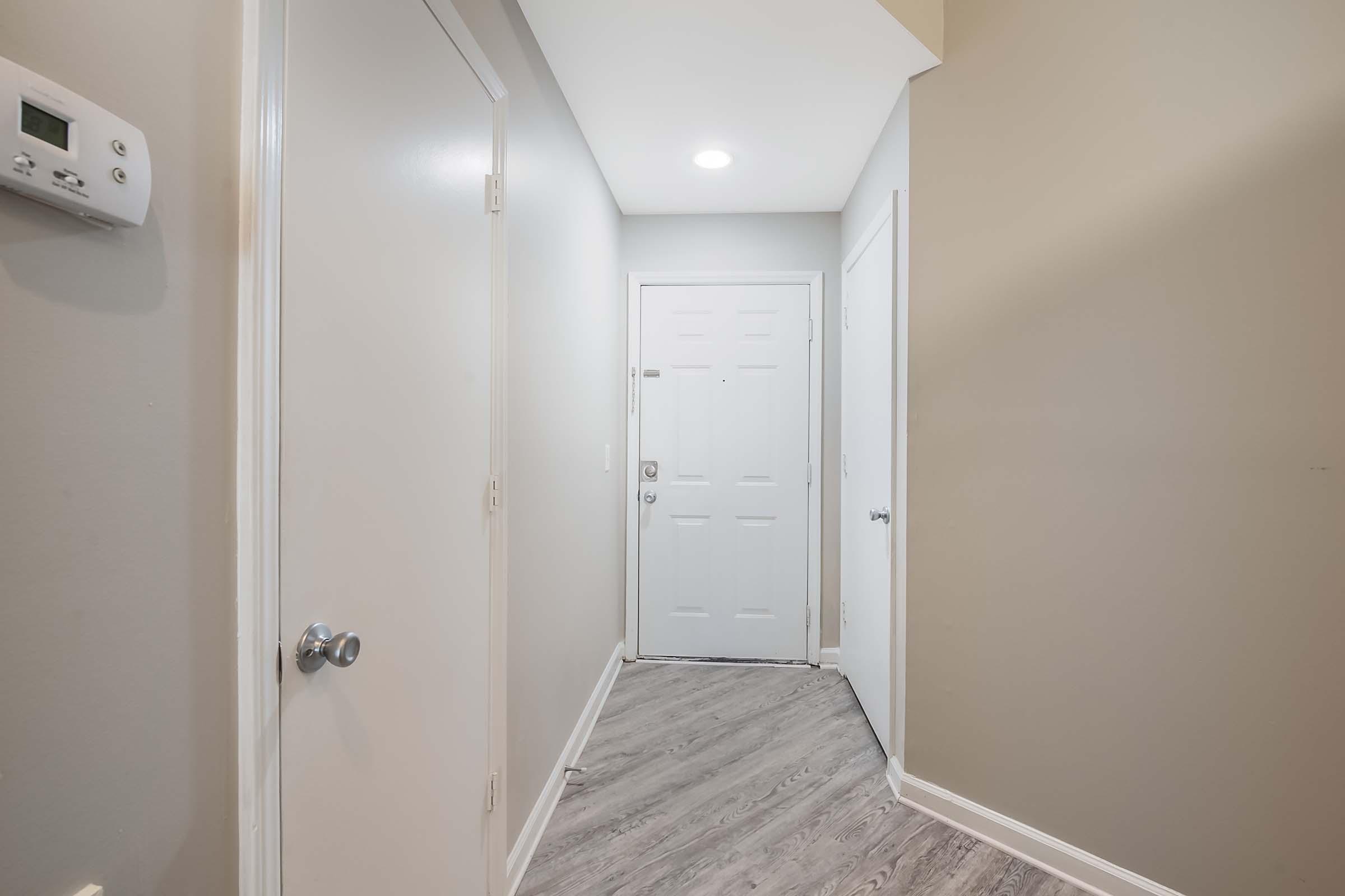 A residential hallway featuring light gray walls, a white front door at the end, and a side door on the right. The floor has a wood-like finish, and there's a wall-mounted thermostat on the left. Natural light comes from an overhead ceiling light, creating a welcoming atmosphere.